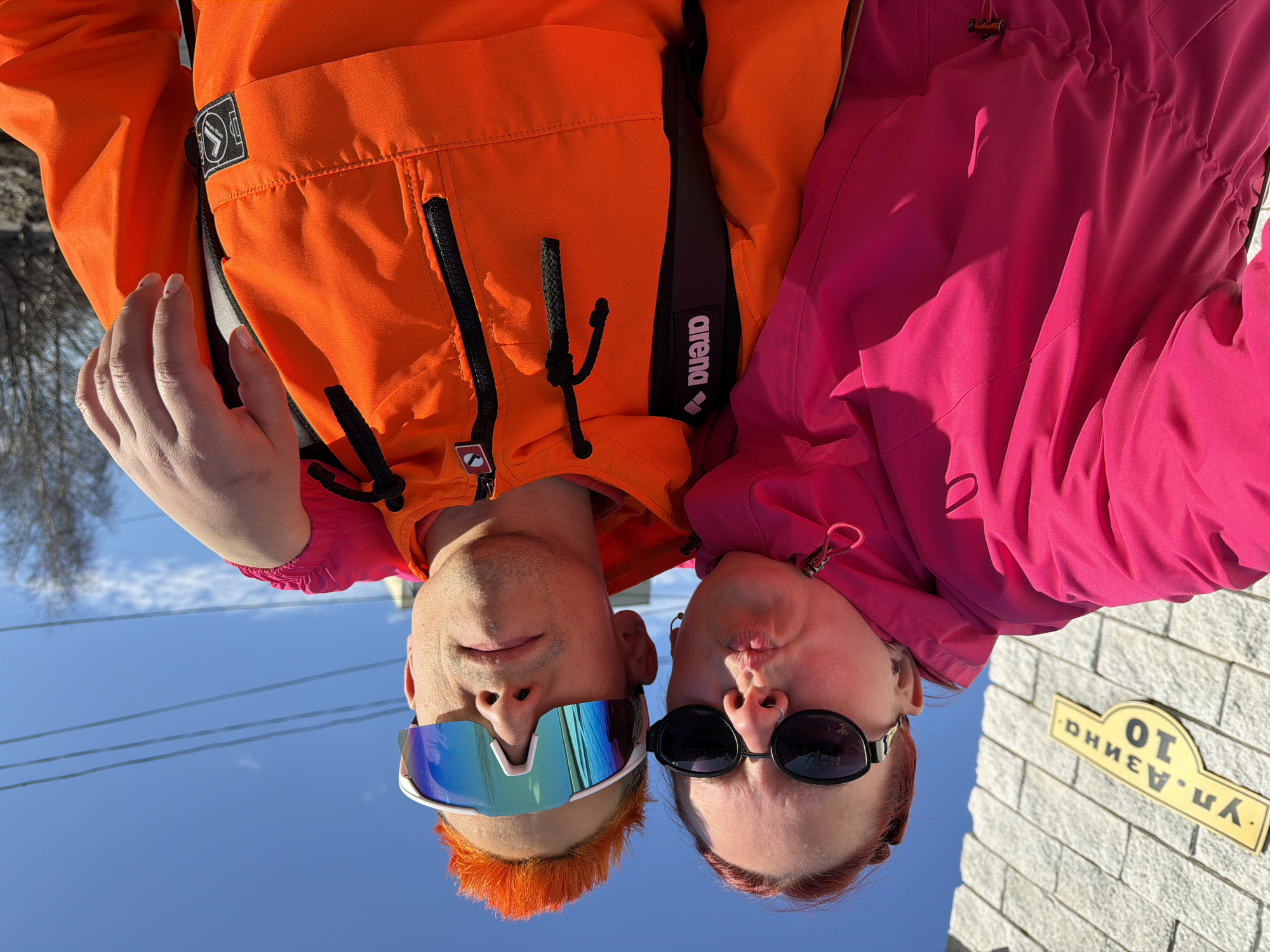 Two individuals, a male and a female, are oriented upside down in the image. The male, on the left, has orange hair and is wearing mirrored sunglasses. He is clad in an orange jacket with black accents. The female, on the right, wears dark sunglasses and a bright pink jacket. They are positioned close together.
In the background, a clear blue sky is visible, along with power lines. A portion of a building is also visible, featuring a street sign. The sign reads "ул. Дзина 10", identifying the location as Dzin Street, building number 10. Based on the street name written in Cyrillic, the location is Chaikovsky, Russia.