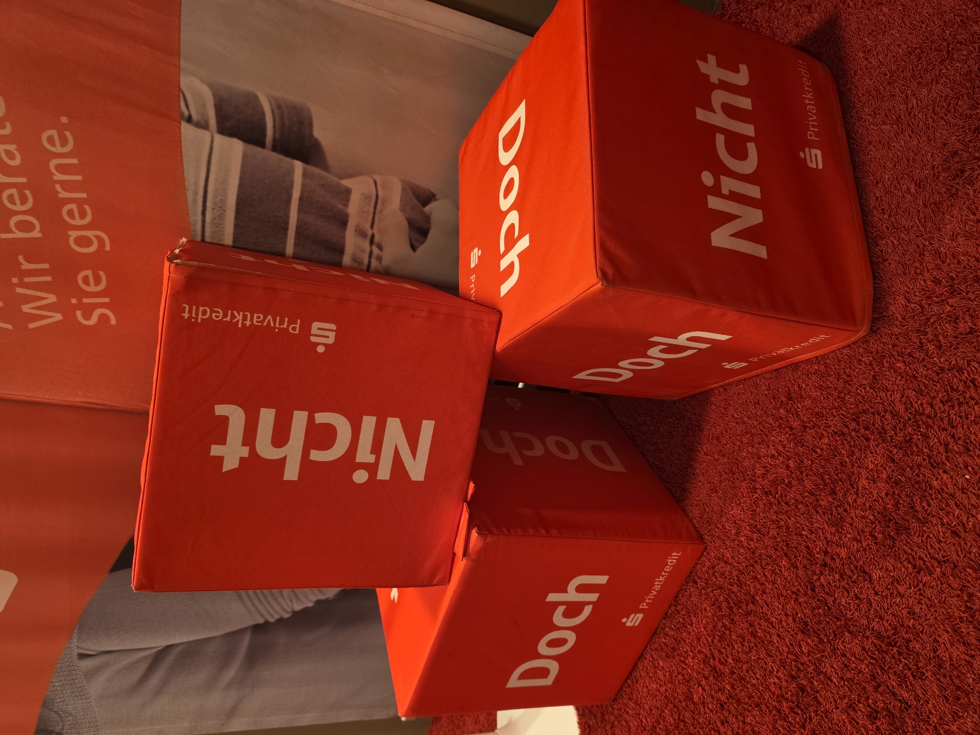 The image depicts three red cube-shaped objects placed on a red carpet. Each cube has white text and a small logo printed on its sides. The text visible includes "Nicht", "Doch", and "Privatkredit". The cubes are stacked and arranged in a way that suggests a display or promotional setup. In the background, a banner with German text is visible. The overall scene appears to be indoors, possibly a promotional event or exhibition. The specific location in Höxter, Germany cannot be determined from the image alone.