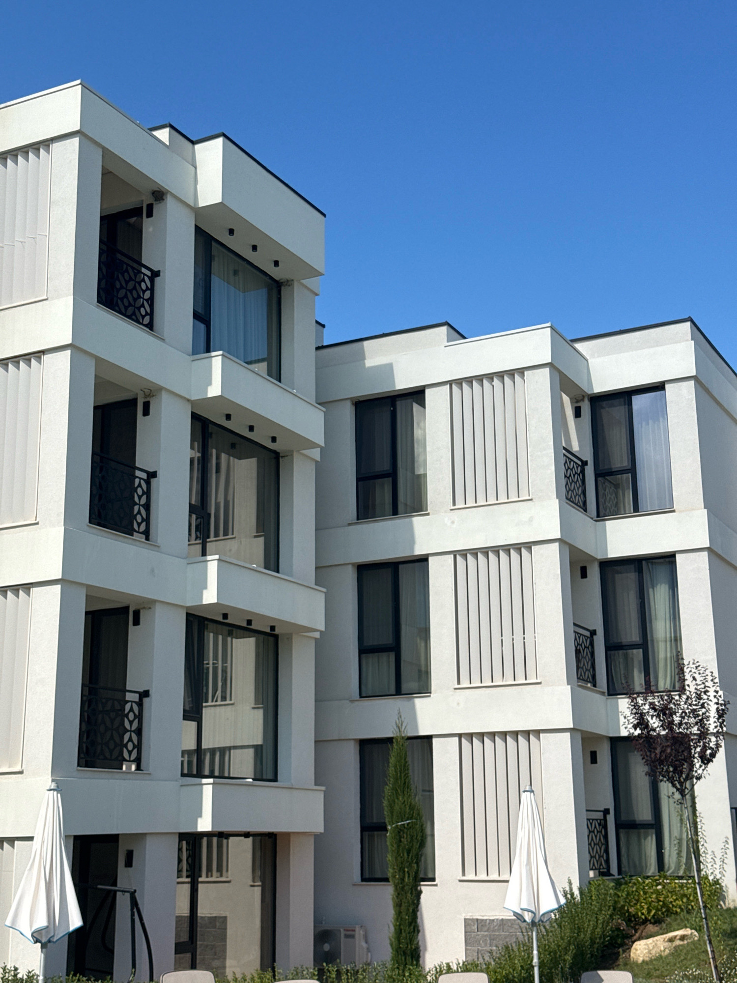 The image depicts a modern, multi-story white building with a clear blue sky as the backdrop. The building features a contemporary architectural style with sharp lines, large windows, and balconies with decorative black railings. Vertical louvers are incorporated into the facade of the building, adding a textured element. In front of the building, two white patio umbrellas are visible, one partially deployed and the other fully open. Lush green bushes and a tall cypress tree are also present, along with a small, dark-leafed tree on the right side of the frame. The scene suggests a sunny day in a resort or residential area, likely in Sozopol, Bulgaria, given the architectural style and the presence of outdoor amenities often found in coastal tourist destinations. There are no people visible in the image.