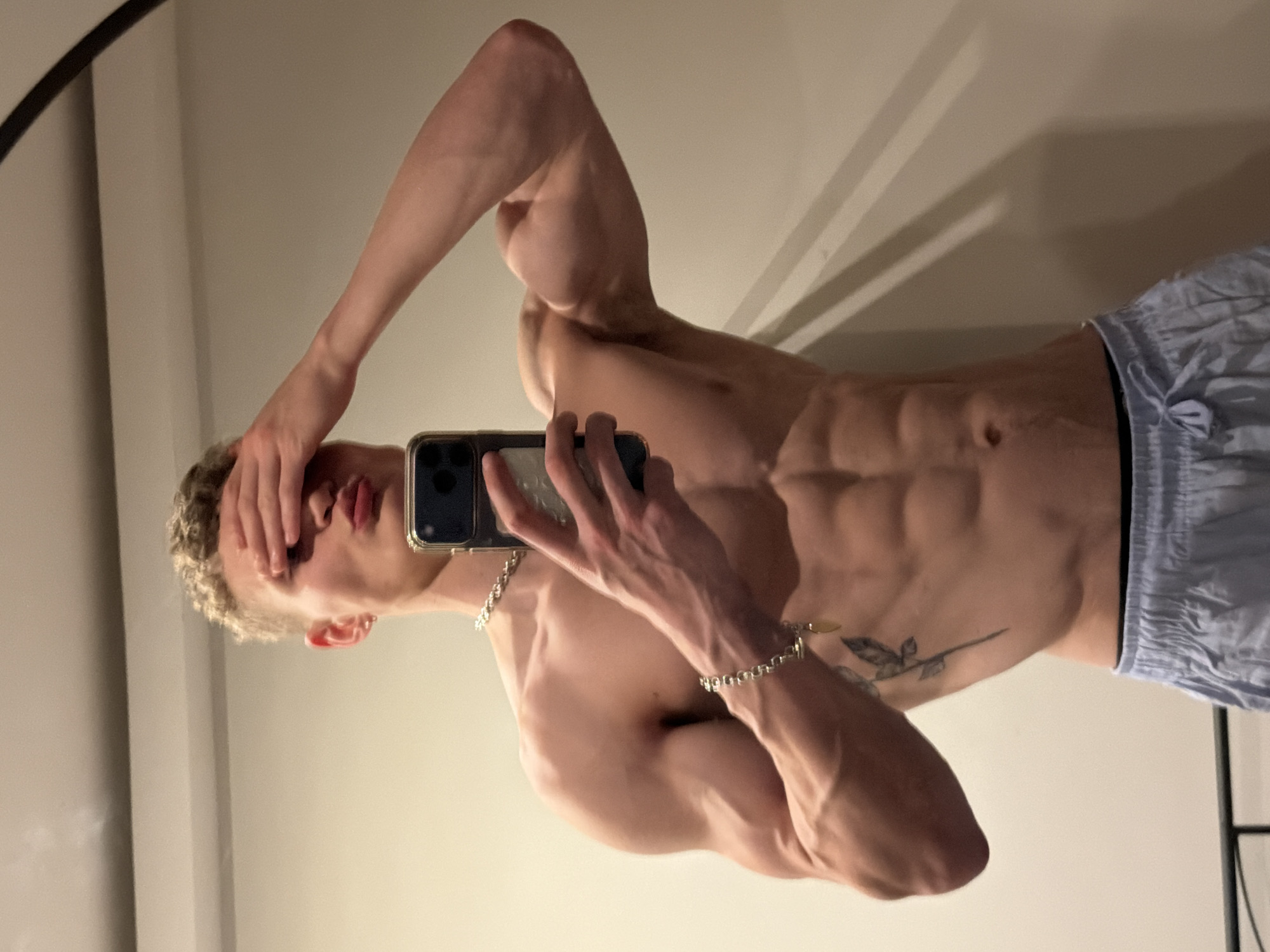 A shirtless, fair-skinned male stands in front of a mirror, holding a smartphone and covering his face with his left hand. He is wearing light blue shorts with a dark waistband. A silver bracelet is visible on his right wrist, and he has a tattoo on his left side. The background consists of plain, light-colored walls with visible shadows. The location cannot be determined.