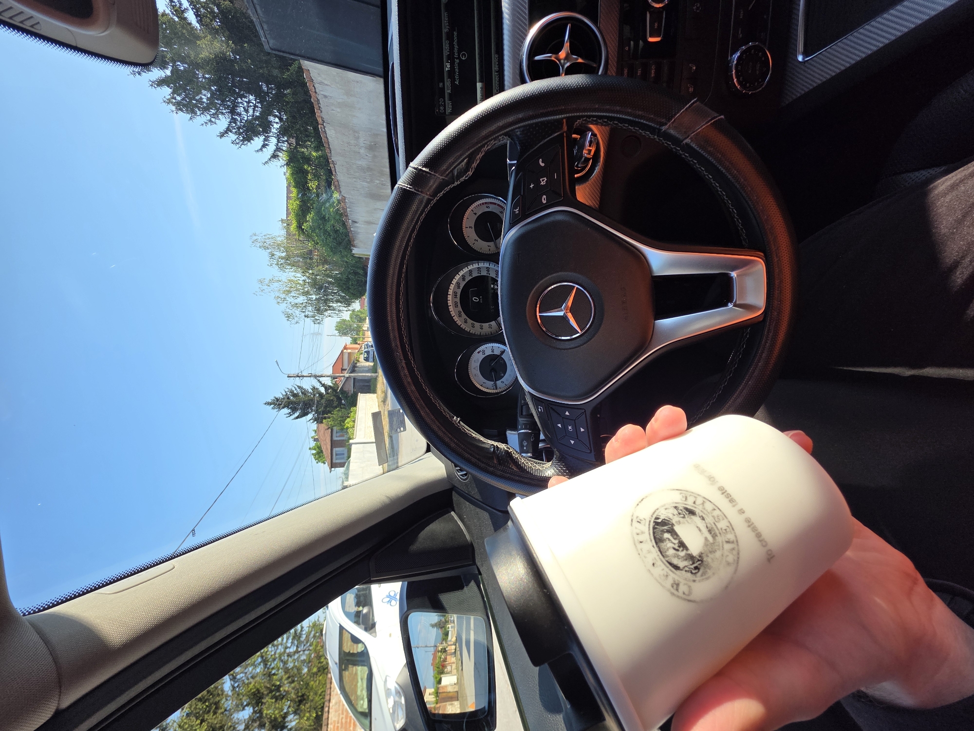 The image shows the interior of a car, focusing on the steering wheel with a Mercedes-Benz logo. A hand is holding a white mug with a "CREATIVE CAFE STYLE" logo printed on it. Through the car window, one can see houses and greenery, an ambulance parked outside, the view corresponds to the city of Yagodovo, Bulgaria. The sky is clear and blue.