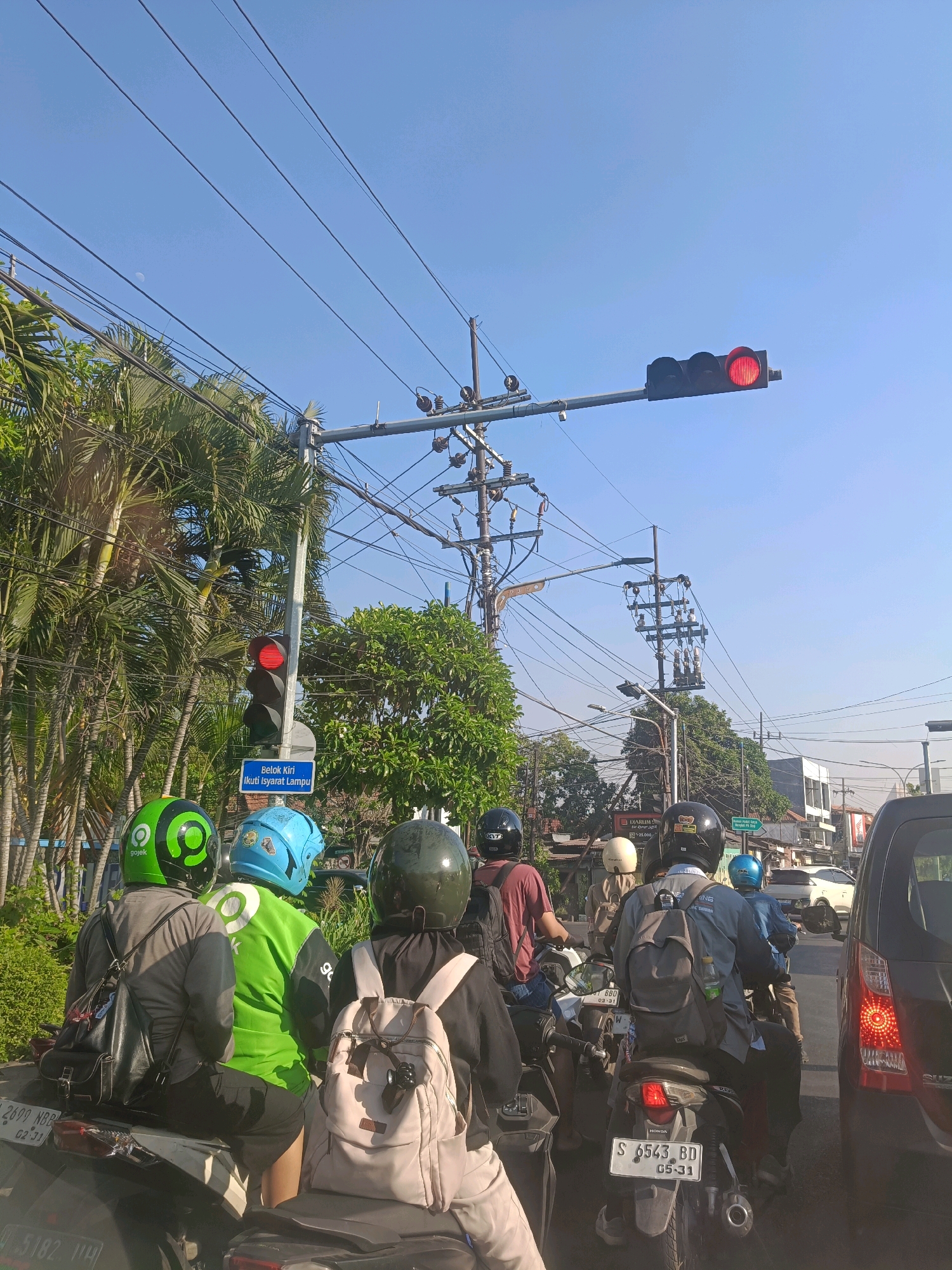 The image depicts a traffic scene in Surabaya, Indonesia, characterized by a bright, clear sky and an abundance of overhead electrical wires. In the foreground, several motorcyclists are stopped at a traffic light, which is illuminated red, indicating a halt. The motorcyclists are wearing helmets of various colors, including green, blue, and black. Some are carrying backpacks. Behind them, more vehicles and motorcyclists are visible, suggesting a congested urban environment. Palm trees and other greenery line the side of the road, contrasting with the man-made infrastructure of power lines and traffic signals. A blue sign is visible on the traffic light pole with text in Indonesian. The overall atmosphere is that of a typical daytime traffic situation in a bustling Indonesian city.