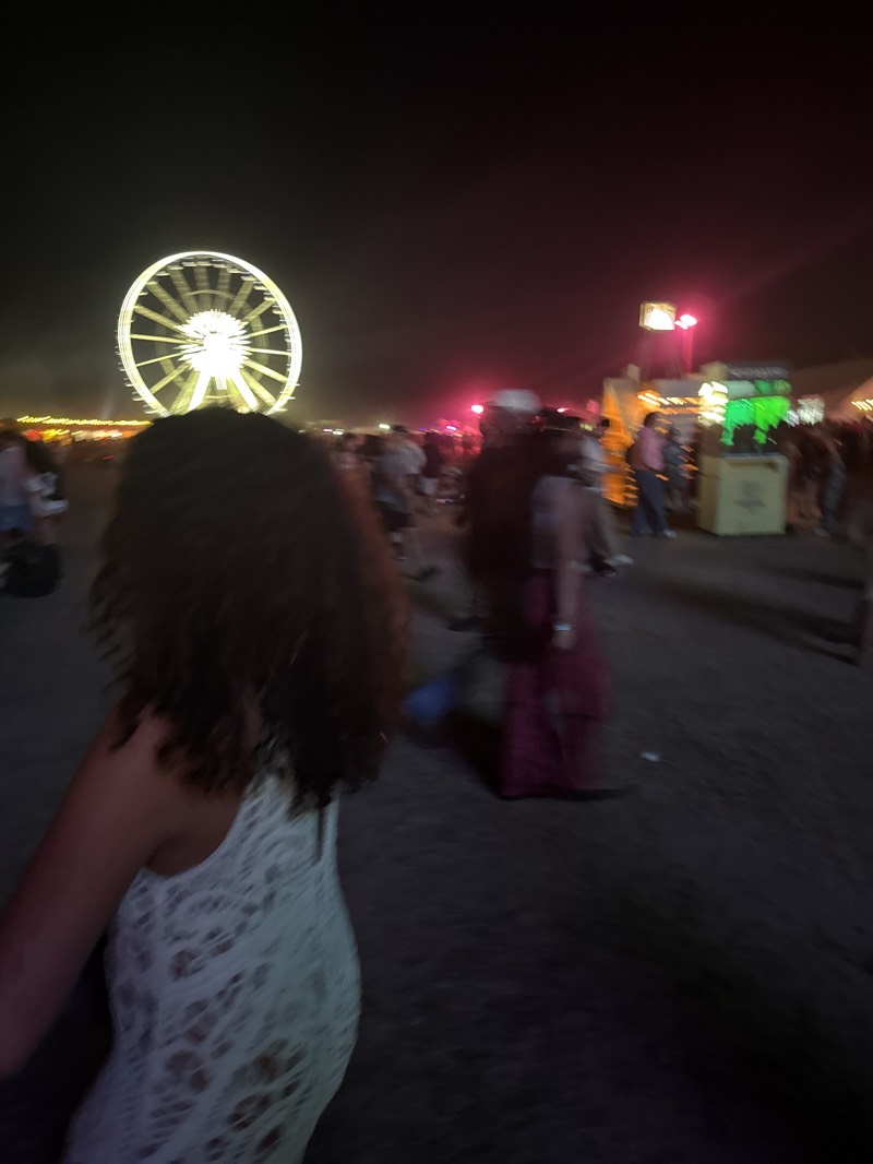 At night, in Indio, United States, a blurry scene depicts a large illuminated Ferris wheel in the background. In the foreground, a woman with brown curly hair, wearing a white dress, is seen from the back. Several people are present in the midground, some standing and some walking. A vendor stand with green and gold signage is to the right. The environment is dark.