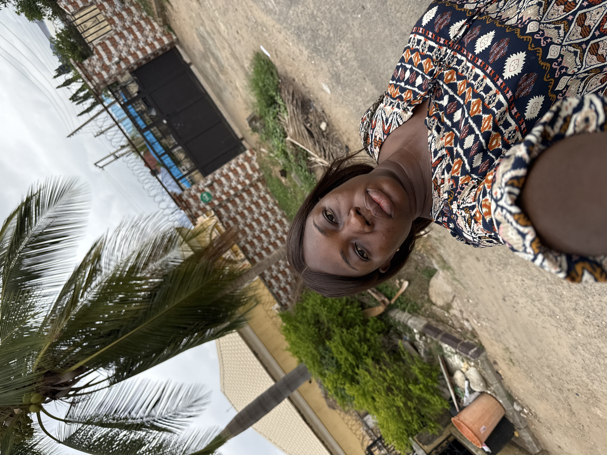 A selfie taken by a dark-skinned woman in Abuja, Nigeria, shows her wearing a patterned dress. The background features a palm tree, a brick and black metal gate secured with barbed wire, and greenery suggesting a residential area. A partially visible beige building, a roadside drain, and a brown plastic bucket are also visible. The ground appears to be a mix of dirt and pavement.