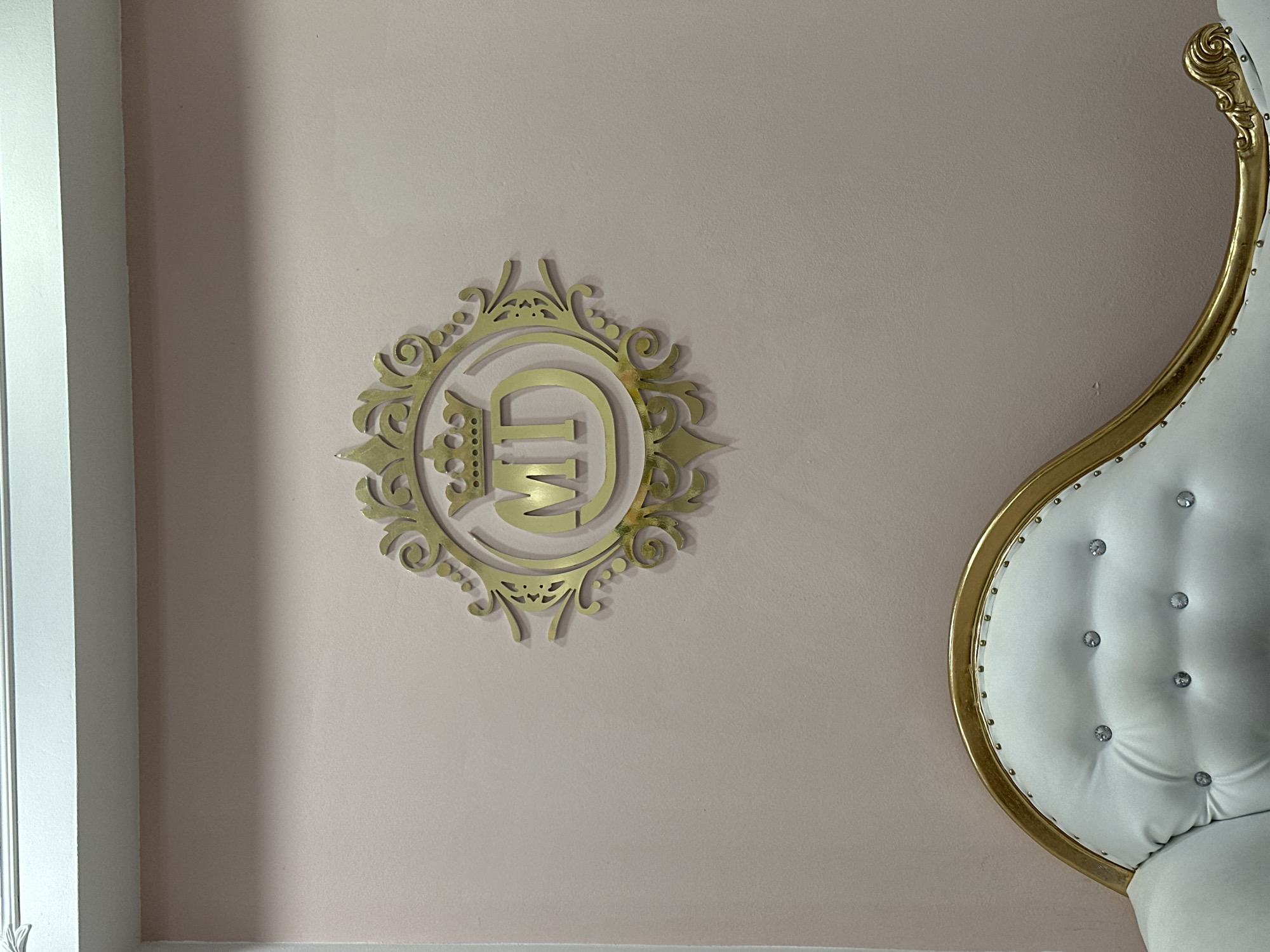 The image shows a close-up of a wall, likely interior, in Sofia, Bulgaria. The wall is light pink and features a decorative, ornate, gold-colored emblem affixed to the center-left. The emblem appears to be a monogram or crest with the letters "MD" inside an elaborate circular frame with floral and scrollwork details. To the right, partially visible, is the top of a white and gold ornate chair. The chair features white upholstery with embedded gems or buttons and a curved gold frame. A white wall frame is seen on the left edge of the photo.