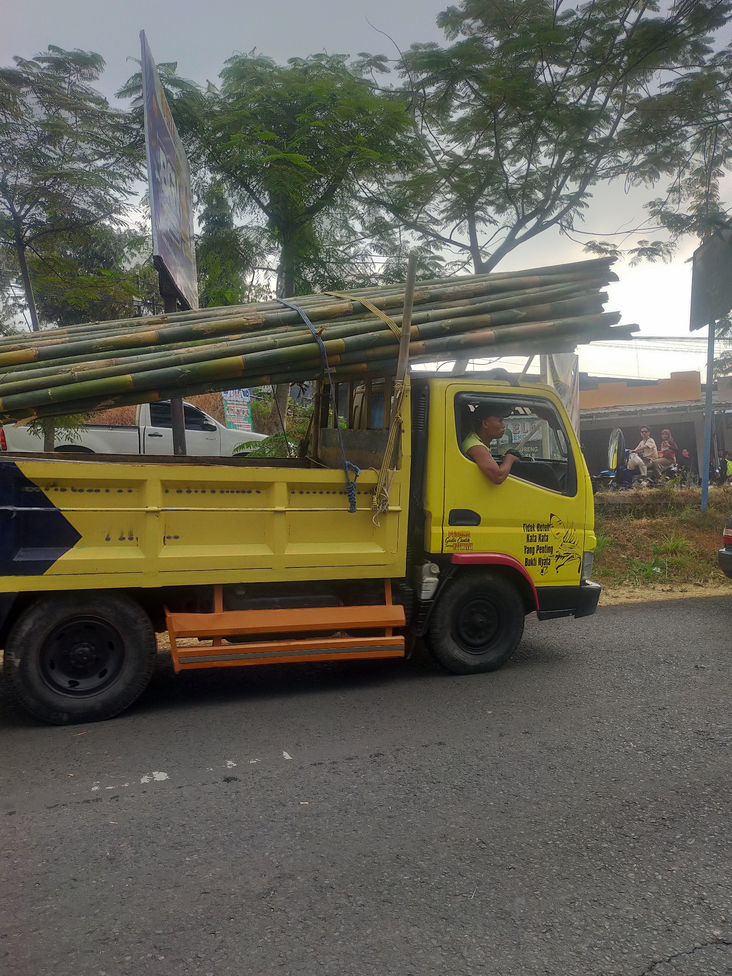 A yellow truck is driving down a street in Tuwel, Indonesia. The truck is loaded with a large quantity of bamboo poles that are tied down with ropes. The truck has a driver in the cab, and there are buildings and trees visible in the background. The sky is overcast.