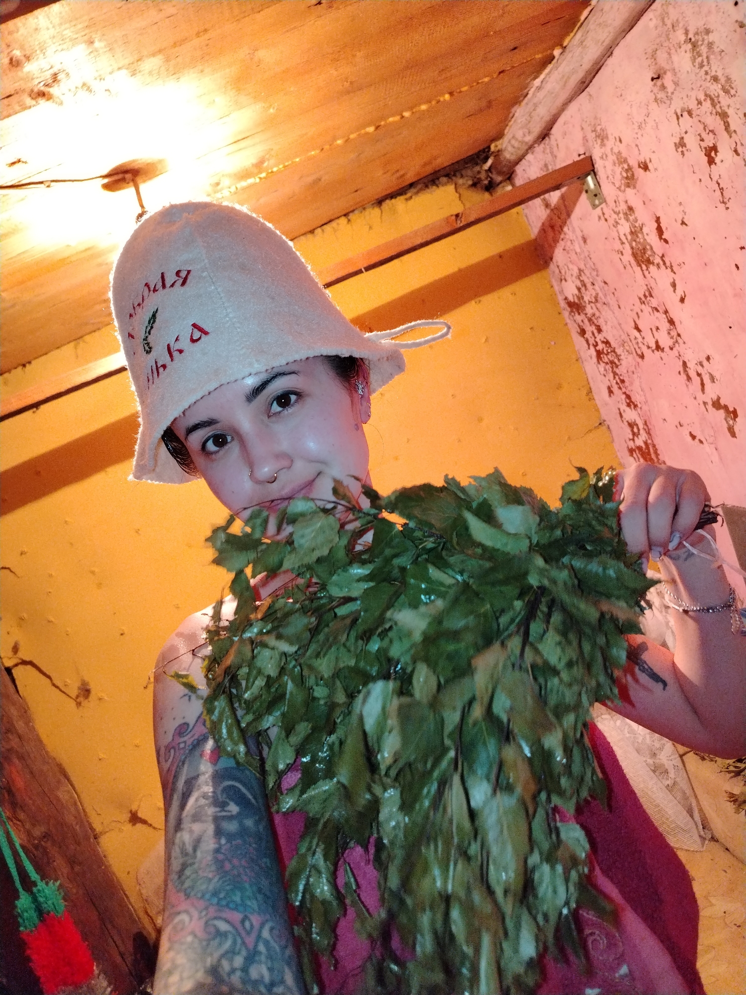 An image taken in what is described as Гожан, Russia, depicts a young adult female posing in a rustic indoor setting, consistent with a traditional banya (sauna). The woman occupies the central and right portions of the frame, looking directly at the camera with a slight smile. She wears a light-colored, possibly felt, sauna hat adorned with Cyrillic script and a small green emblem, and a nose ring in her left nostril. Her right shoulder and arm are visible, covered with extensive, colorful tattoos. She is wrapped in a reddish-pink garment and holds a large, wet bundle of green leaves, consistent with a *venik* (banya broom), in front of her with her left hand. A thin bracelet is visible on her left wrist.
The background features a wooden plank ceiling, from which a single incandescent light bulb hangs, emitting a strong warm light and causing a lens flare near the top center. The wall to the right is a faded reddish-pink, showing signs of significant wear, texture, and possibly peeling material. A horizontal wooden beam and a metal bracket are attached to this wall. The wall to the left is a solid orange-yellow. A small, red knitted object with green tassels hangs from a wooden element in the lower left foreground. The overall lighting is warm and artificial, suggesting an enclosed, humid environment.