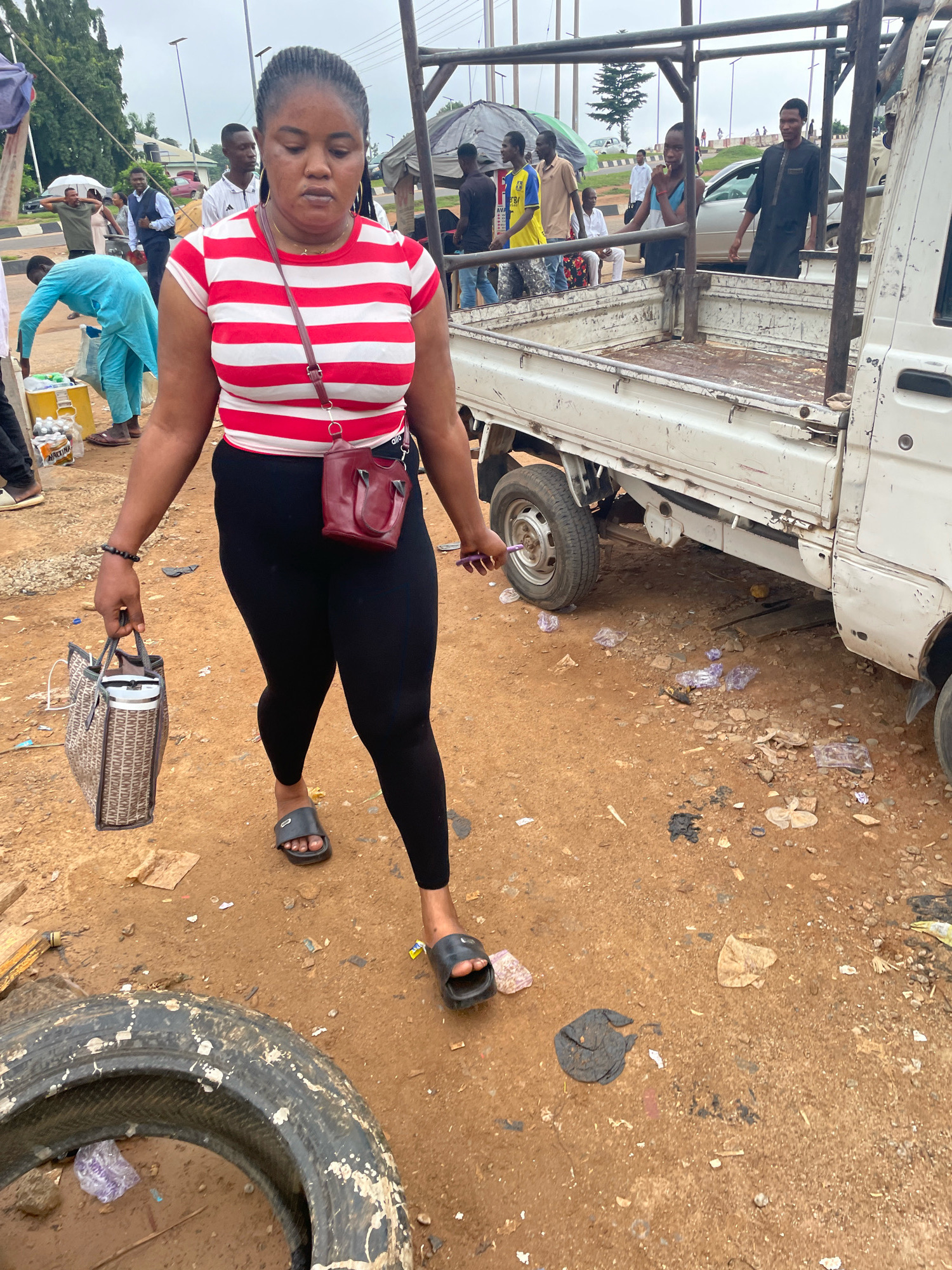 A young Black woman with braided hair is walking on a dirt road in Abuja, Nigeria. She is wearing a red and white striped t-shirt, black leggings, and black flip-flops. She is carrying a tan patterned tote bag in her left hand and a small, burgundy purse is slung across her body. In the background, a white pickup truck is parked, and other people are visible. The ground is covered with dirt, debris, and some scattered garbage. There is a large, worn tire lying on the ground in the foreground.
