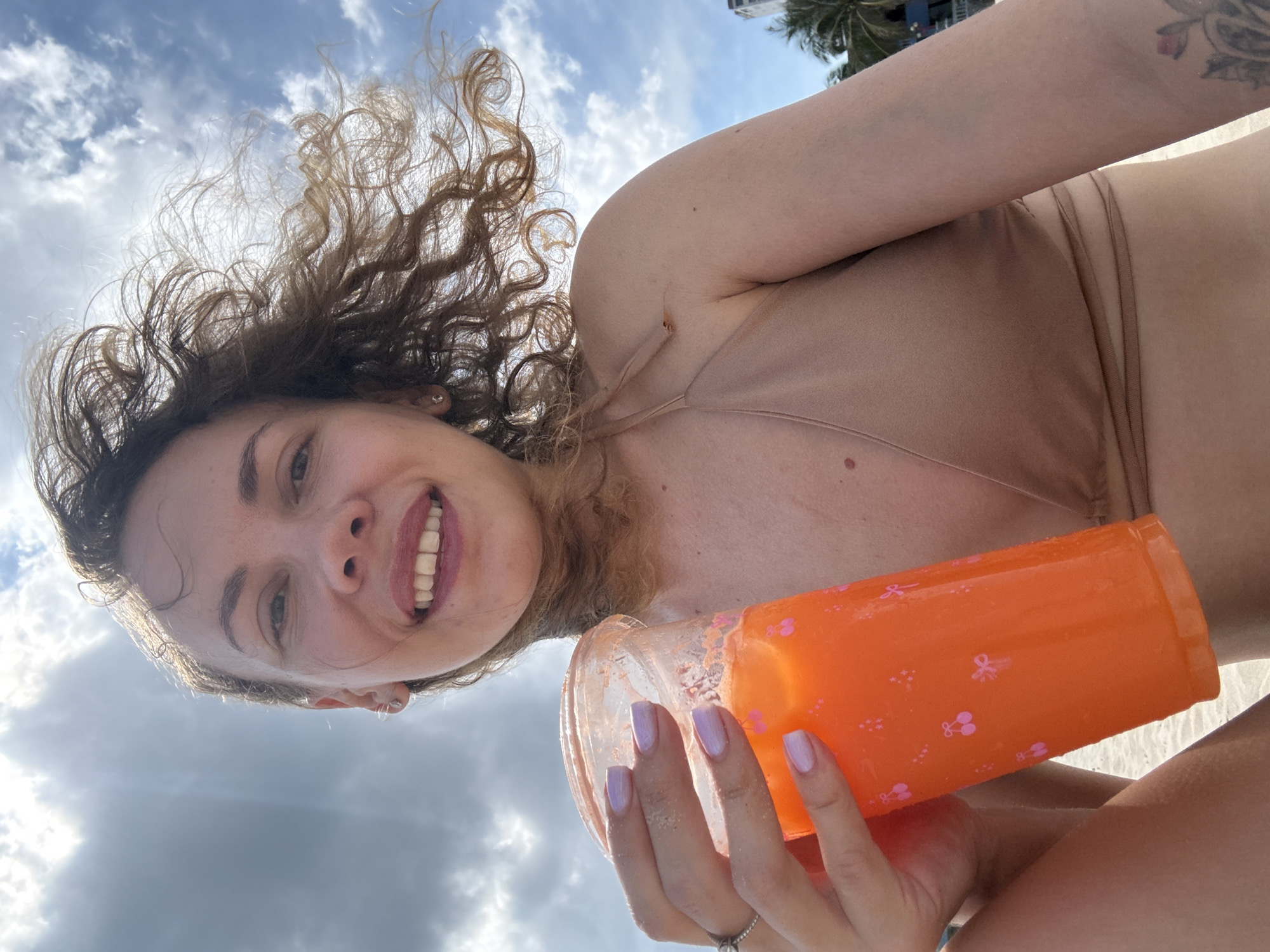A woman with curly brown hair and fair skin smiles for a selfie on a beach. She is wearing a beige bikini top and holding an orange plastic cup filled with an orange beverage and ice. The cup has small pink cherry and bow designs. Her nails are painted a light purple. Her left hand, with a ring on her ring finger, is holding the cup. The background shows a bright blue sky with scattered clouds and sunlight. Some palm trees and buildings are visible in the distance. The woman appears to be on a beach, possibly in Mũi Né, Vietnam, given the tropical foliage and ocean setting.