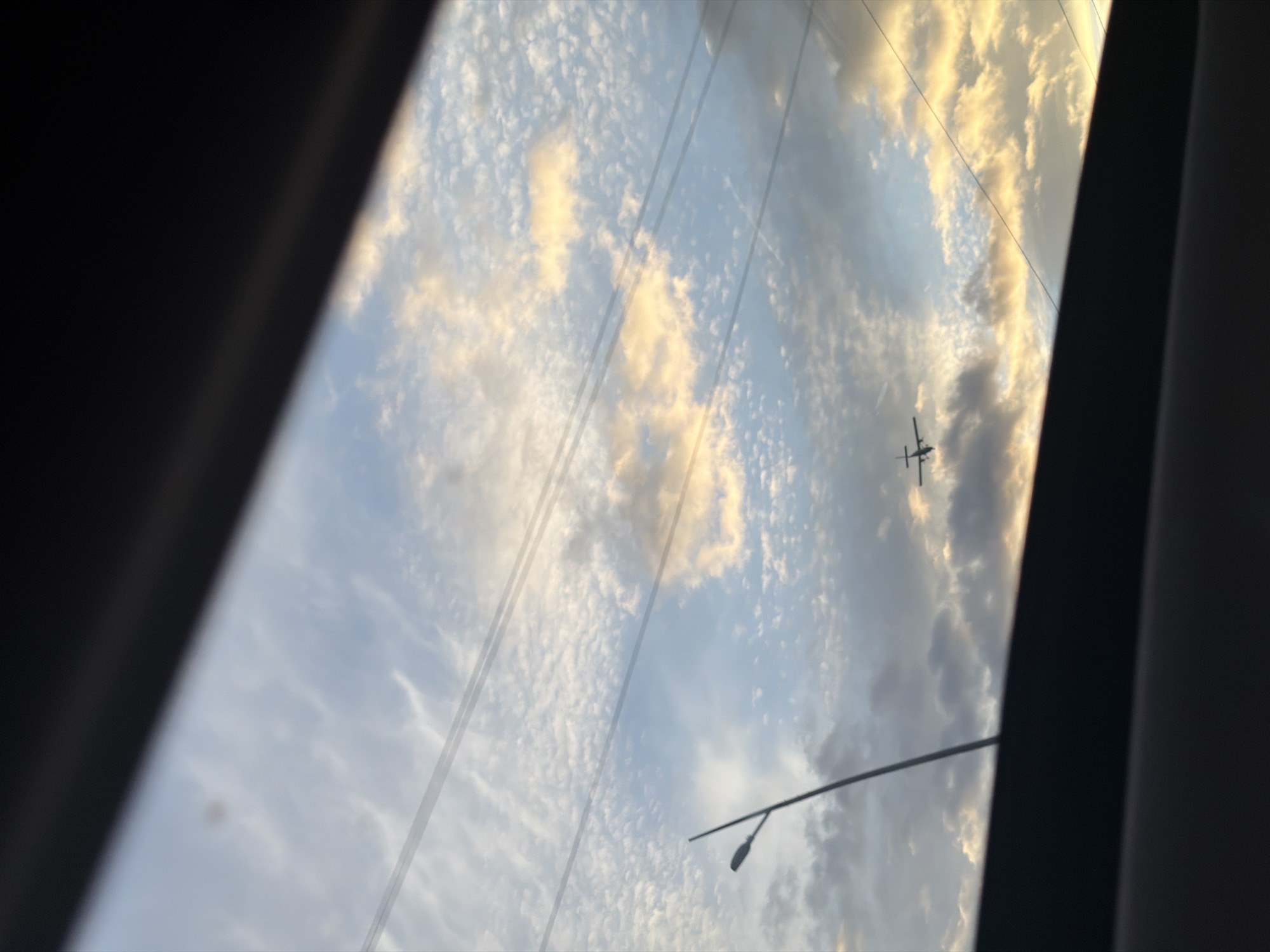 The image shows a view of the sky through a window, likely from inside a vehicle or building. The sky is filled with clouds, and the lighting suggests it might be dusk or dawn. A small plane is visible in the sky. There are also power lines or cables running across the frame. A street light is also visible. The location, Krasnoyarsk, Russia, cannot be determined from the information provided in the image.