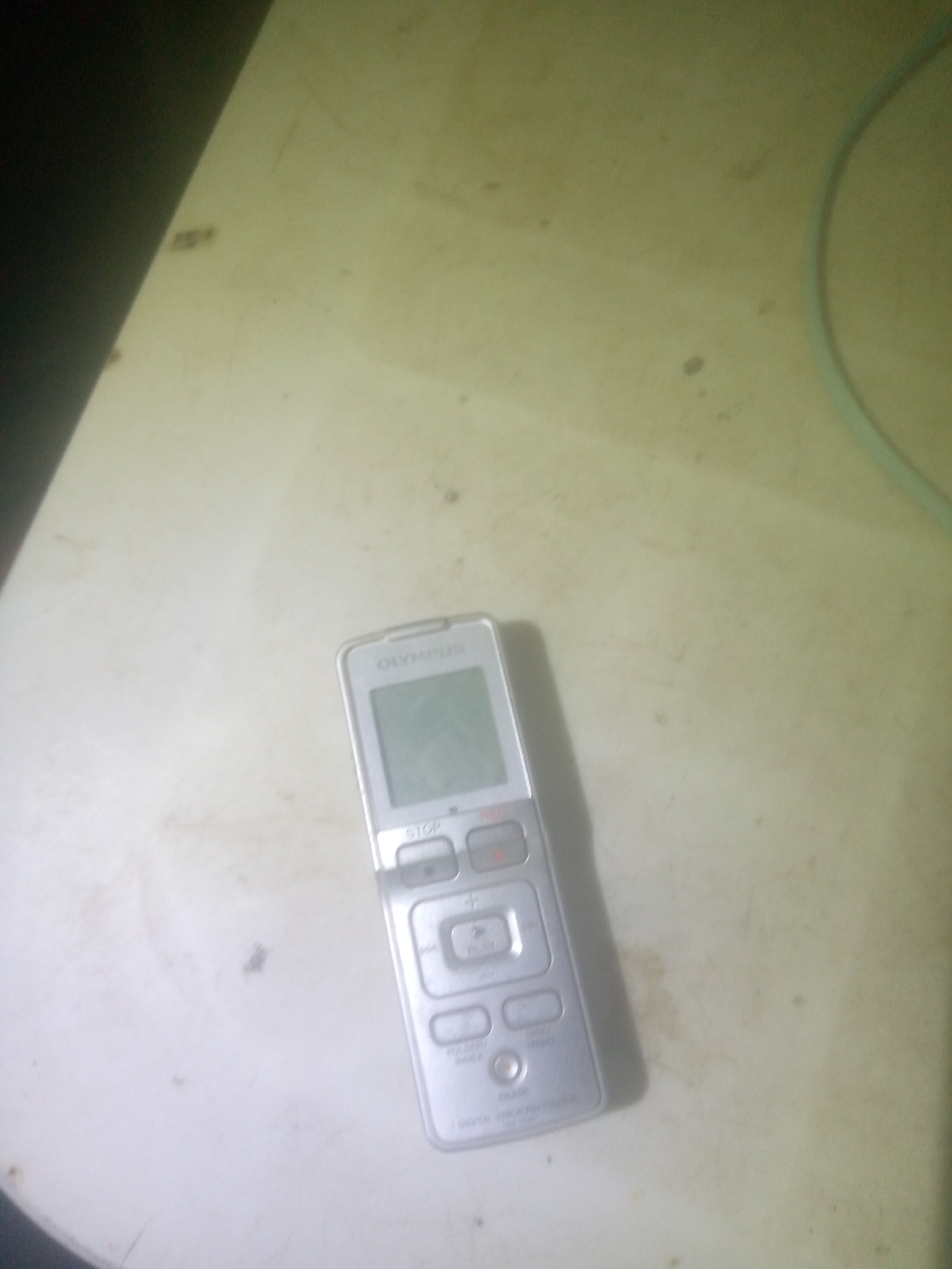 A silver Olympus digital voice recorder lies on a light-colored, textured surface. The recorder has a screen, a red record button, and several other buttons including "STOP", "PLAY", and "FOLDER/INDEX". The surface appears to be a tabletop or desk with some smudges and a faint circular discoloration. A light blue cord curves into the frame from the upper right corner, partially obscured by a dark shadow on the left side of the image.