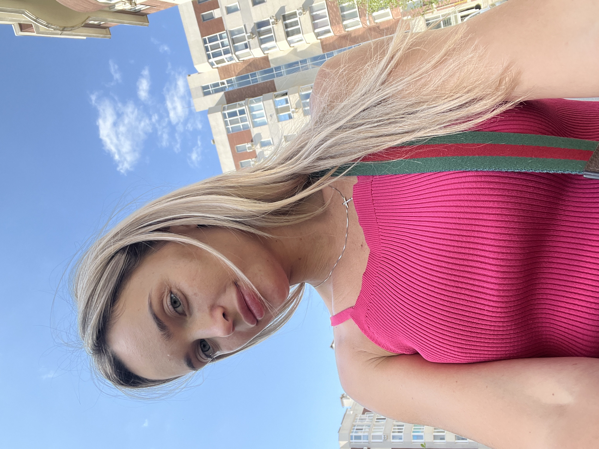 A blonde woman is captured in a close-up, low-angle selfie outdoors against a clear blue sky with scattered white clouds. She is wearing a bright pink ribbed tank top and a necklace with a small cross pendant. A green and red striped strap is visible across her shoulder. In the background, modern apartment buildings with light-colored facades and numerous windows are visible, suggesting an urban or suburban setting. The specific location is identified as Kryzhanivka, Ukraine. The lighting indicates it is daytime, likely sunny.