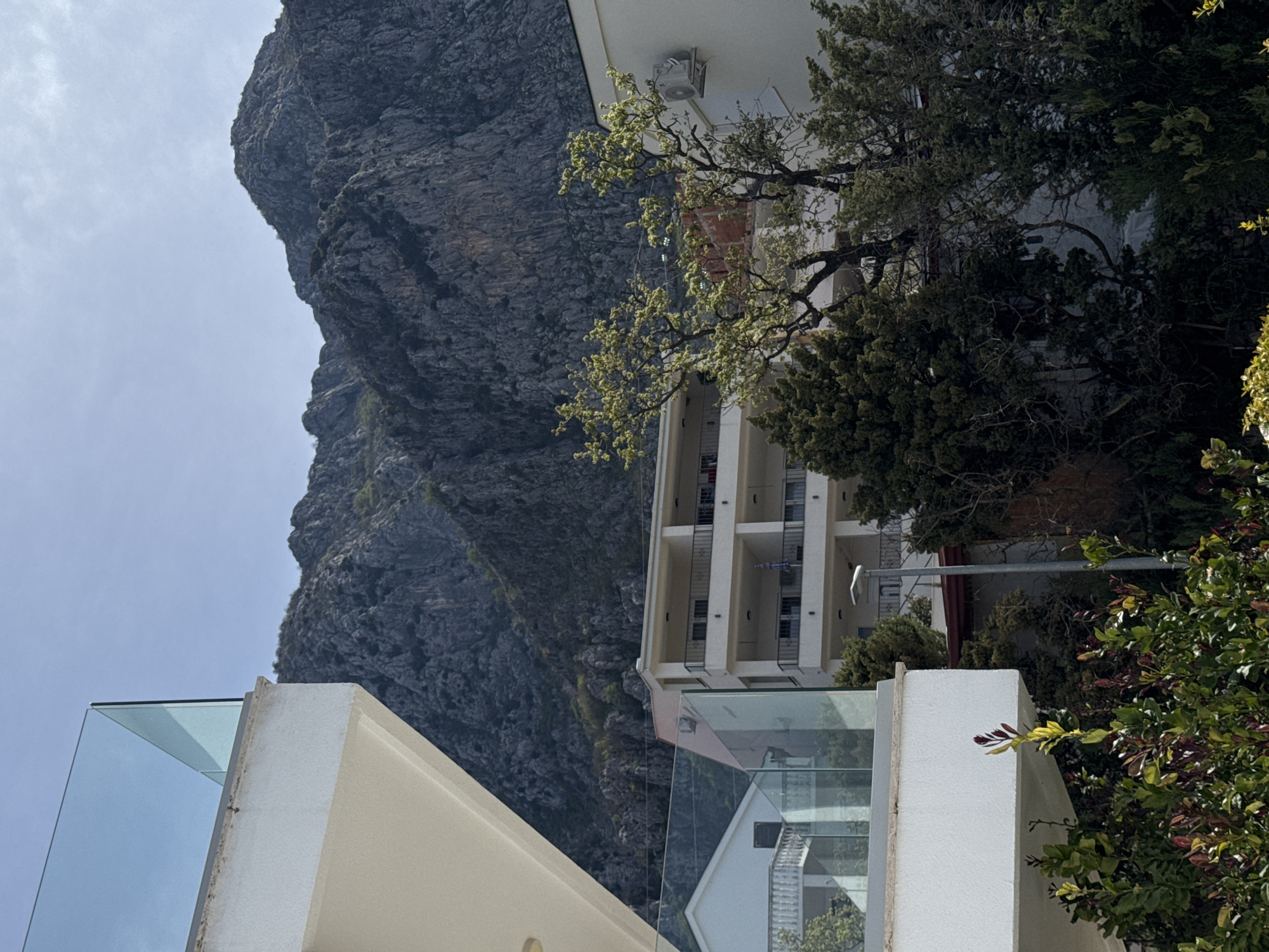 The image presents a view in Blizikuće, Montenegro, showcasing a rugged mountain terrain meeting modern architecture. A light-colored building with multiple balconies stands prominently, partially obscured by foliage. The presence of a glass-paneled structure and adjacent concrete elements suggests contemporary construction. The mountain backdrop, with its steep, rocky face, contrasts sharply with the built environment. The sky is overcast, casting a soft light over the scene.