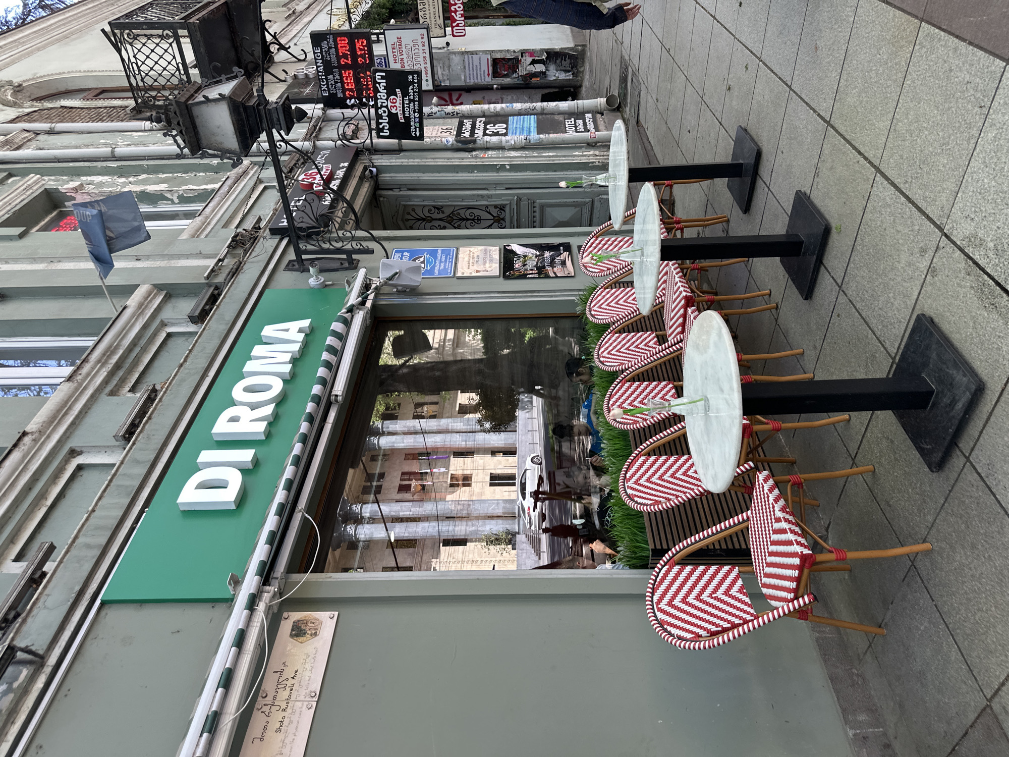 The image depicts the exterior of the "DI ROMA" cafe in Tbilisi, Georgia. The cafe has a green awning and a sign with white lettering. Outside, there are several small, round tables with red and white woven chairs. A plaque with Georgian text is displayed on the building's facade. The reflection in the cafe's windows shows a building with columns. A currency exchange rate sign, an additional hotel advertising, and various other signs are visible on the adjacent buildings. A person's leg is visible in the top right corner.