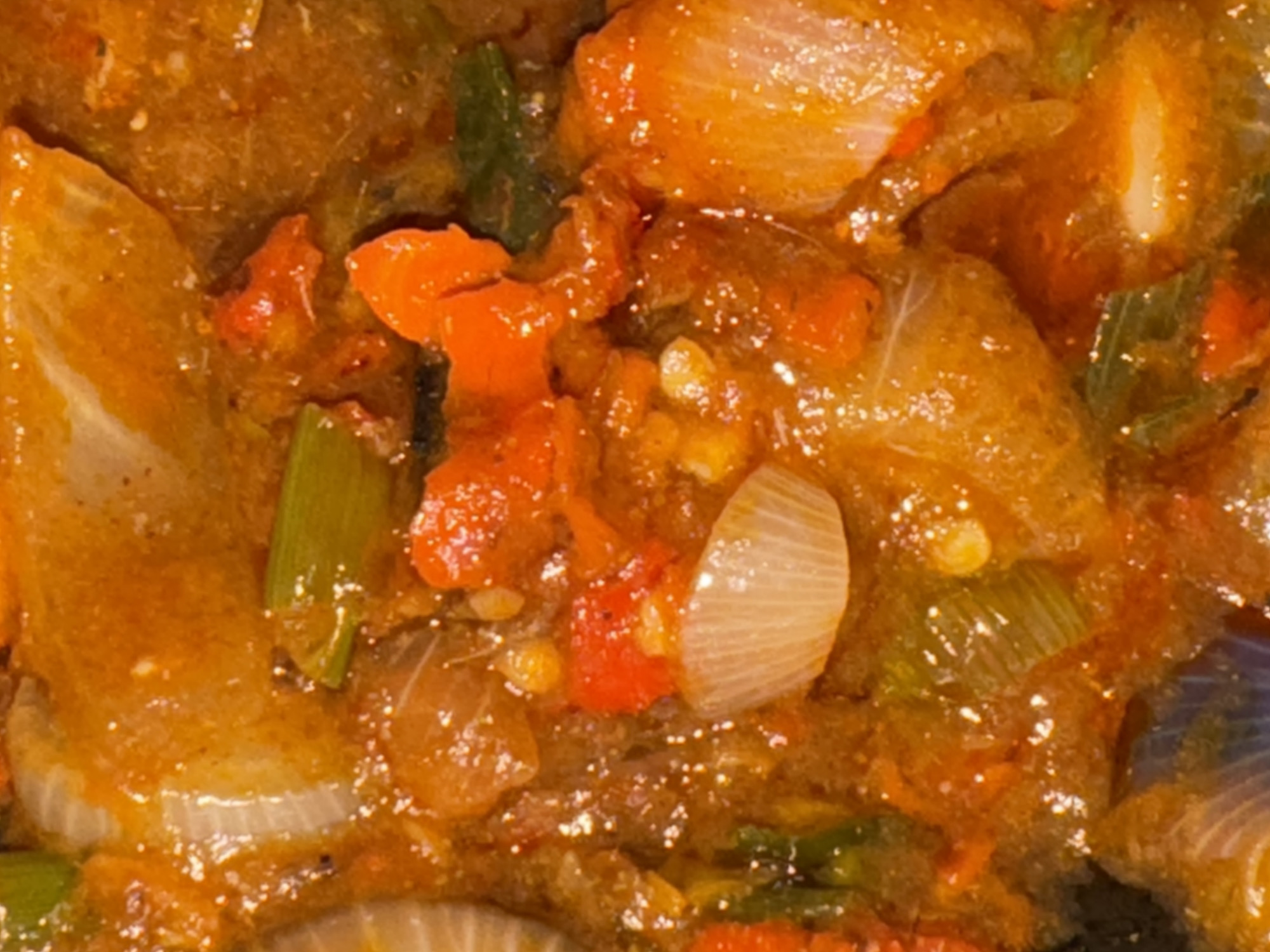 This close-up image displays a vibrant, cooked dish, likely a stew or sauce, characterized by its rich orange-brown color and glossy, saucy consistency. The primary visible components are generously sized, translucent slices of white onion, bright red diced pieces that appear to be tomatoes or red bell peppers, and distinct pieces of chopped green herbs or vegetables, possibly spring onions. These ingredients are thoroughly incorporated into the thick, simmering sauce. Small specks, possibly spices or seasonings, are visible throughout the liquid, contributing to the texture. The food appears well-cooked and soft, suggesting a hearty and flavorful preparation. There are no people, specific activities, or visible text in the image. Given the location context of Kurunduma, Nigeria, the dish likely represents a local or regional cuisine.