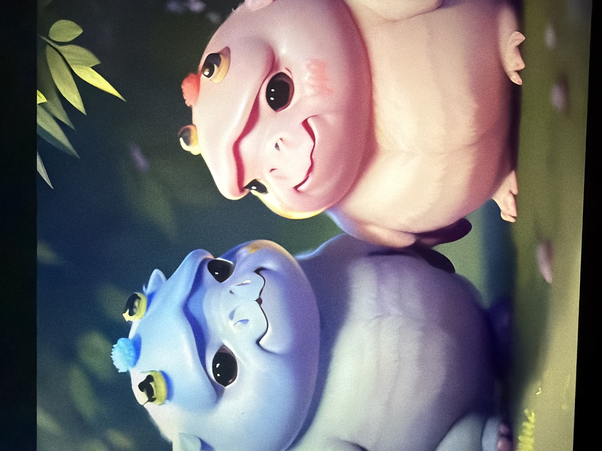This is a digital illustration featuring two cartoon-like creatures. The creature on the left is light blue with large, dark eyes and a small smile. It has yellow and black protrusions on its head, along with a blue tuft. The creature on the right is pink, also with large dark eyes and a smile, and has red and yellow protrusions on its head. Both creatures appear plump and have short limbs. They are positioned close together in a grassy, outdoor environment. The background is dark and out of focus, with hints of green leaves and dark foliage at the top. The lighting suggests a soft, ambient glow. There is no indication of this scene being located in Kyiv, Ukraine, or any specific city.