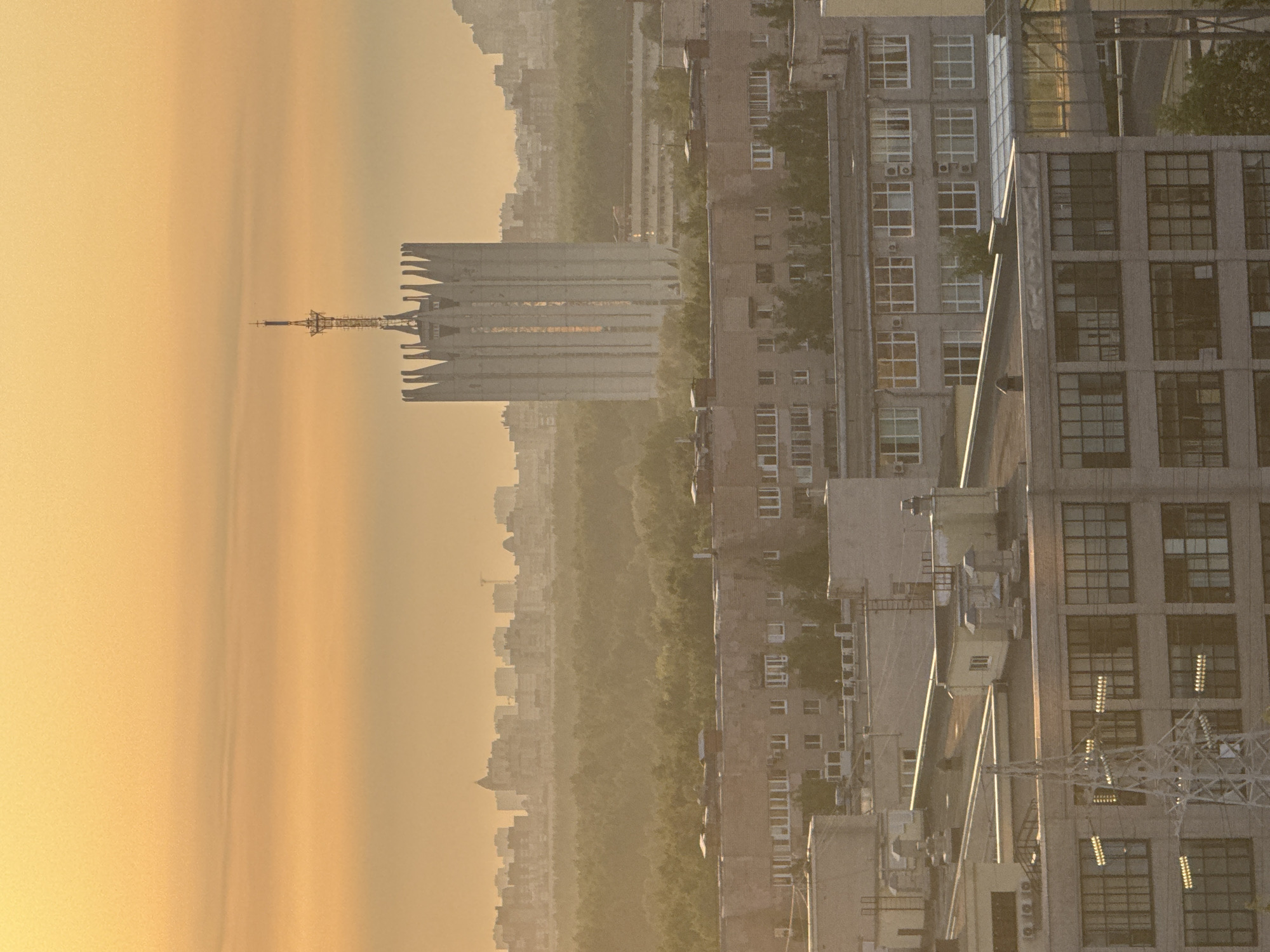 The image shows a cityscape under a hazy yellow sky, likely at sunrise or sunset. Prominent in the distance is a large, distinctive Soviet-era building with a tall radio tower atop it. Closer to the viewer, there are several apartment buildings with visible windows and some air conditioning units. A network of electrical towers and power lines cuts across the lower right corner of the image. Based on the architectural style and tower, the location is likely the city of Sinyavino, Russia, with the distinctive building being a communications or broadcasting center. There are no visible people. The environment suggests a densely populated urban area with significant atmospheric haze.