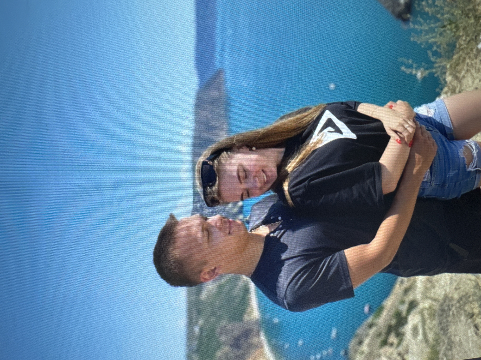 A young couple is embracing outdoors with a scenic blue ocean and cliffs in the background. The man, on the left, has short brown hair and is wearing a dark blue t-shirt with a necklace. The woman, on the right, has long blonde hair, sunglasses perched on her head, and is wearing a black t-shirt with a white logo and denim shorts. They are both looking downwards, and the woman's hands are clasped over the man's on her stomach. The environment appears to be a coastal cliffside with sparse vegetation. The sky is clear and blue.