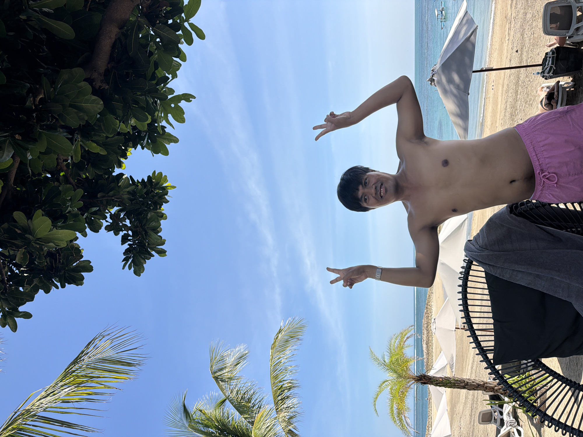 A young Asian man, shirtless and wearing pink swim trunks, stands on a sandy beach in Kuta, Indonesia, making a peace sign with both hands. Palm trees, umbrellas, and lounge chairs dot the beach, with the turquoise ocean and a clear blue sky serving as the backdrop. The man is smiling and appears to be posing for the camera. The image captures a relaxed vacation atmosphere.