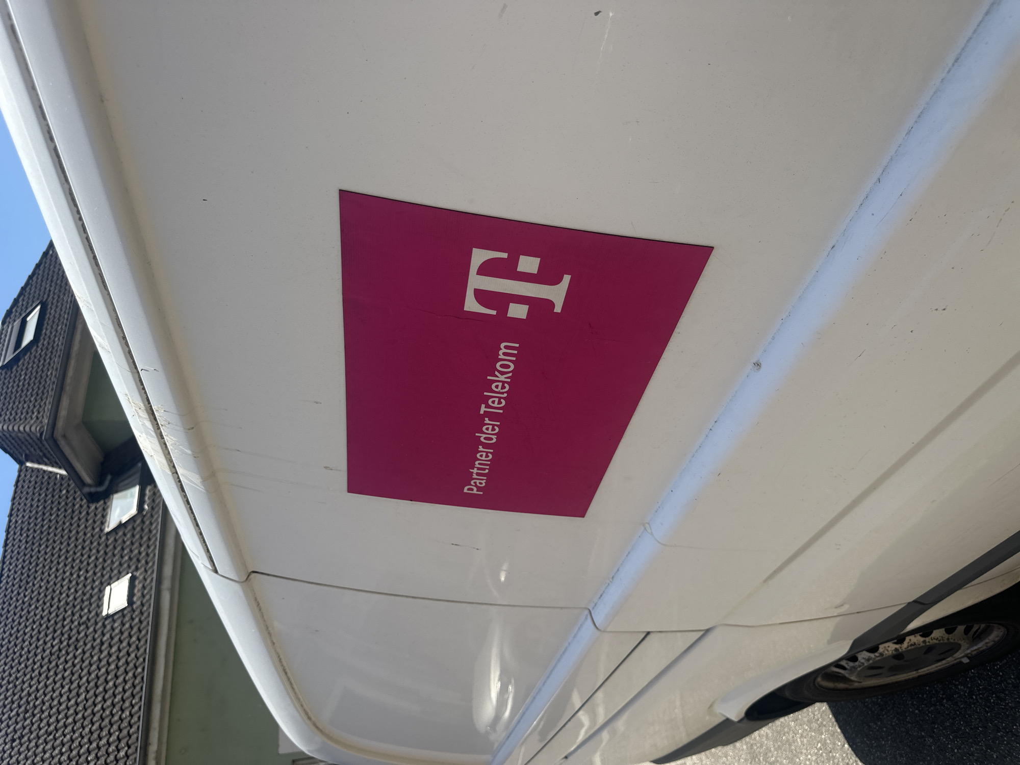 An outdoor, eye-level, medium shot shows the side of a white van with a magenta sticker on the right side. The sticker has white text that reads "Partner der Telekom" and the white Telekom logo. The van is parked on an asphalt surface next to a house with a dark-tiled roof and green siding. The sky is blue with some white clouds.