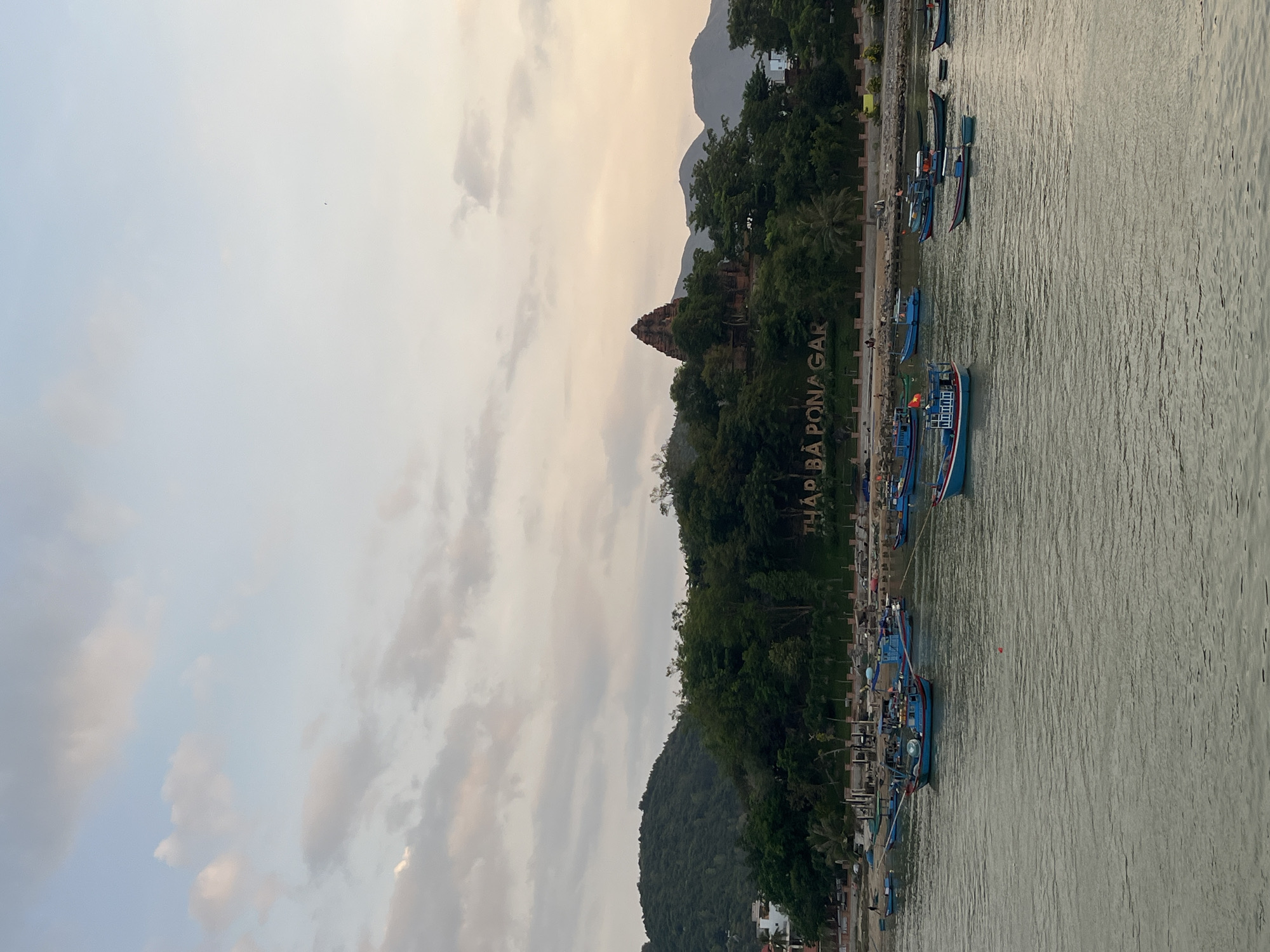 The image shows a view of Tháp Bà Ponagar, a Cham temple tower complex in Nha Trang, Vietnam. It is located in the city of Bac Nha Trang Ward. The temple is situated atop a hill, with the text "THÁP BÀ PONAGAR" spelled out in large letters on the hillside. A body of water, likely the Cai River, runs along the base of the hill, with several blue fishing boats docked along the shore. The sky is cloudy.