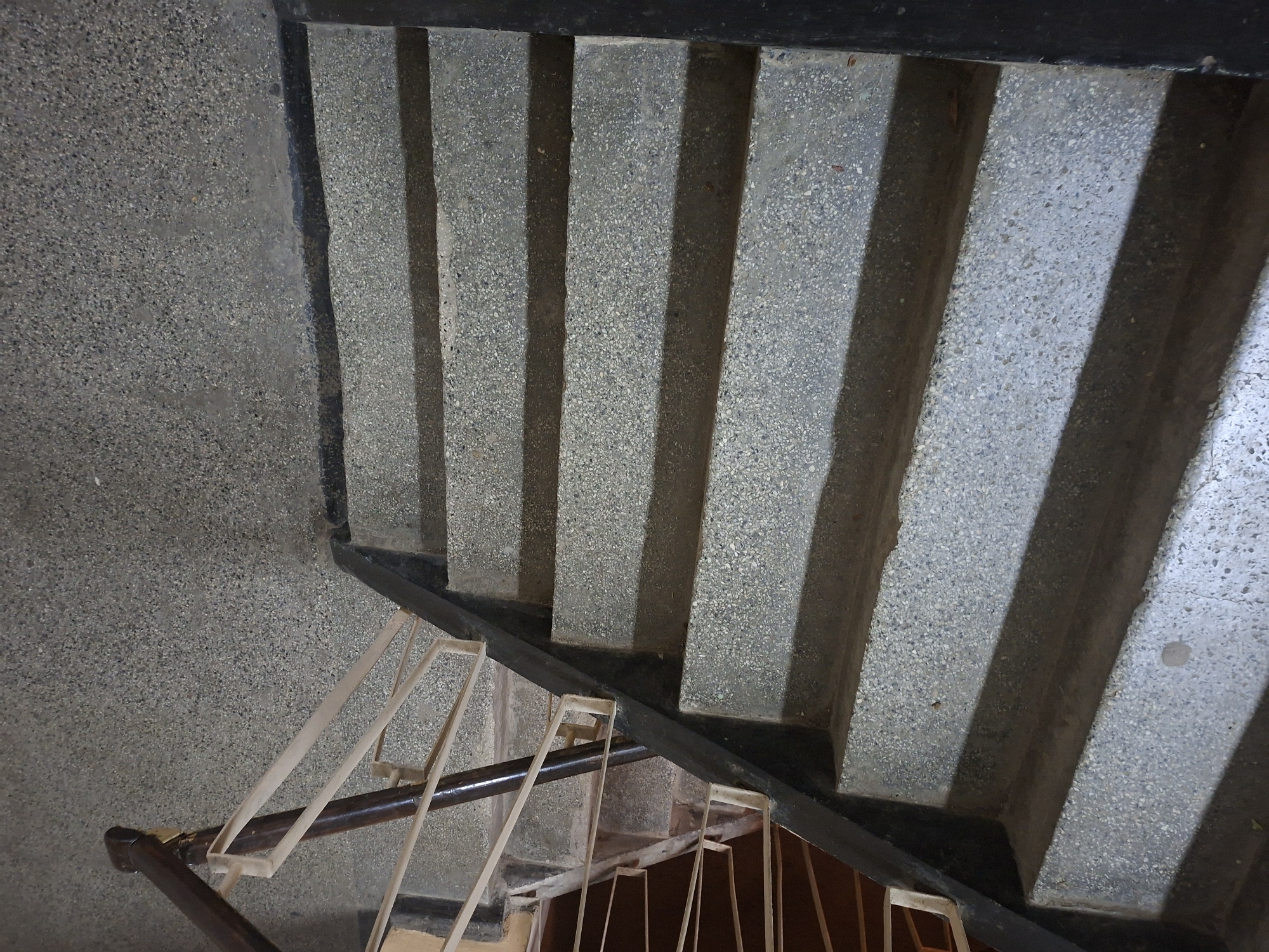 The image provides a top-down view of an indoor staircase, likely located in Pleven, Bulgaria. The stairs and the adjacent wall are constructed from a light grey aggregate material, characterized by a speckled, granular texture reminiscent of terrazzo or exposed aggregate concrete. Each stair tread and riser displays this consistent material.
Prominent, parallel shadows are cast diagonally across the stairs, emphasizing the depth and structure of each step. These sharp shadows suggest a direct light source, likely daylight entering from a window or skylight, indicating the photograph was taken during daytime.
In the lower half of the frame, a dark metal railing with a brown wooden handrail is visible, following the descent of the stairs. The railing features decorative, light-colored metal balusters arranged in geometric, rectangular patterns. The architecture appears robust and functional, characteristic of a public or residential building. There are no people present, and no visible text.