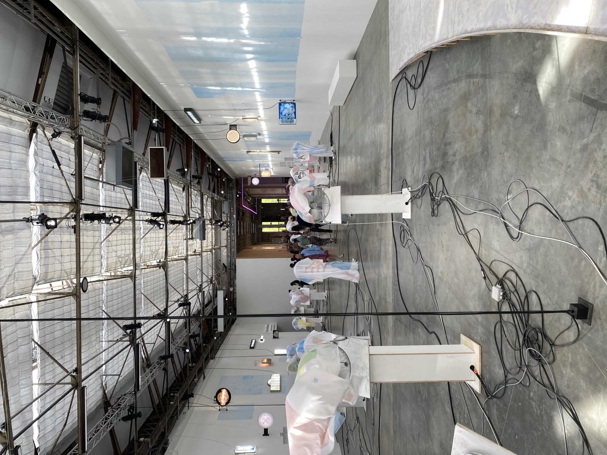 The image captures an expansive indoor space, likely an exhibition hall or industrial building, transformed for an art installation or event. The perspective looks down a long aisle, with the left wall featuring an intricate metal truss structure adorned with numerous spotlights and lighting fixtures. Large, translucent white fabric panels are draped over this structure.
Along the central aisle, a series of white pedestals are arranged, each supporting an electric fan. Draped over these fans are lightweight, translucent fabric pieces, often featuring pastel colors like pink, blue, and white, with subtle patterns. The fans appear to be animating these fabrics, creating dynamic, flowing forms. A tangle of black and white electrical cables lies scattered across the concrete floor, leading to the various installations.
In the background, a line of people, mostly seen from behind, are observing the installations. They are casually dressed, suggesting an informal public event. The ceiling features a distinctive light blue and white painted design, resembling a cloudy sky, providing a contrasting backdrop to the industrial architecture. A large, curved white wall section is visible on the right.
One sign within the exhibition clearly displays the text "City & Humans," with further, less legible text beneath it, possibly an exhibition title or description. The overall atmosphere suggests a contemplative or immersive artistic experience. The setting is an indoor event in Paris, France.
