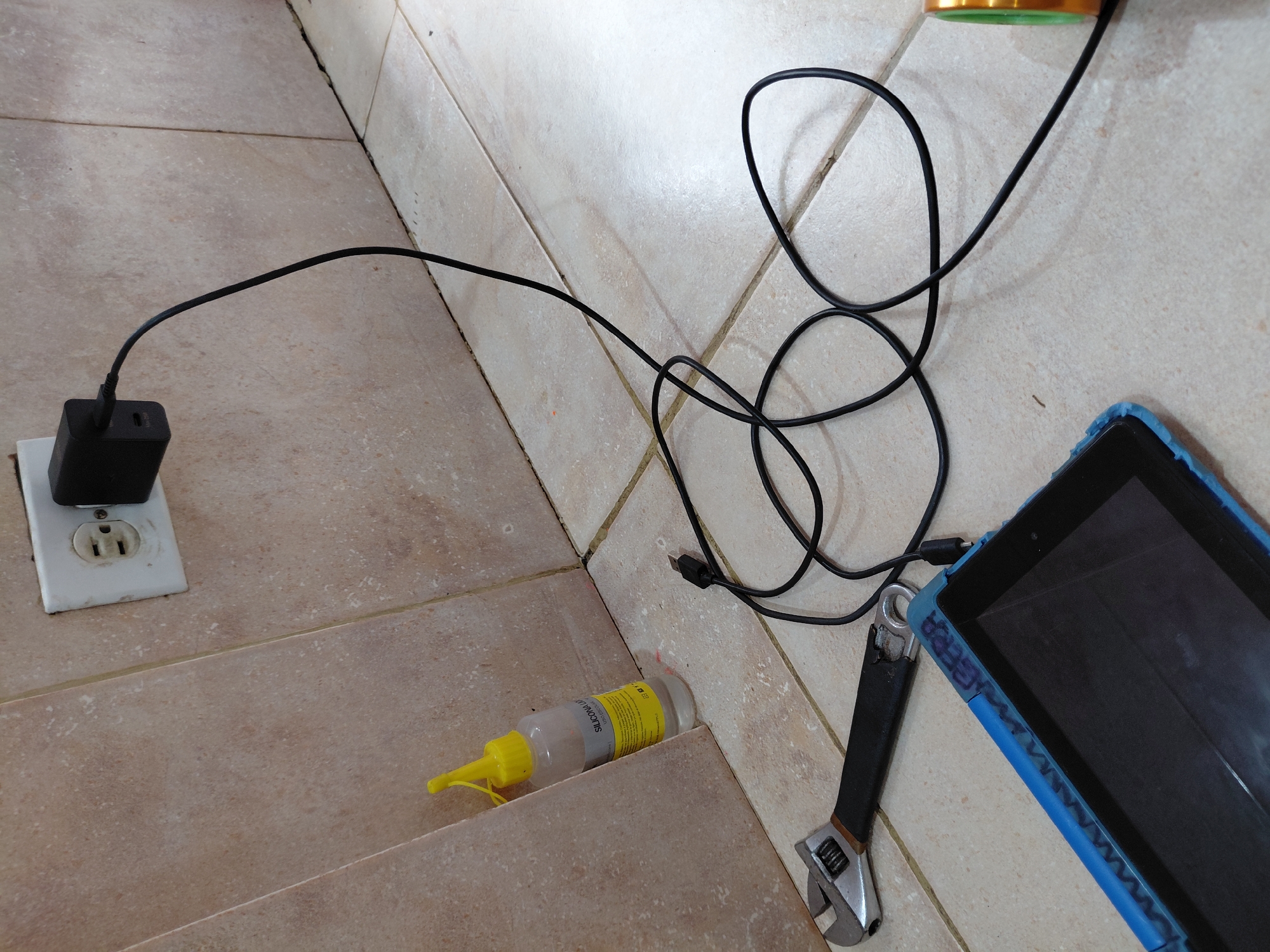 The image captures an indoor scene in Santa Ana, Venezuela, displaying various objects arranged on a light brown tiled floor. In the foreground on the left, a black power adapter is plugged into a white wall outlet. A black charging cable emerges from the adapter, snaking across the tiled surface towards the right.
The cable's far end connects to a black tablet encased in a vibrant blue protective cover, positioned towards the bottom right of the frame. Lying next to the tablet is an adjustable wrench with a silver head and a black-and-silver handle. Below the coiled charging cable, slightly to the left of the center, rests a clear plastic bottle with a bright yellow pointed cap. The label on the bottle clearly reads "SILICONA LÍQUIDA" (Liquid Silicone), along with some smaller, unreadable text. A small part of a gold-colored object with a green rim is visible in the upper right corner.
The primary activity depicted is the charging of the tablet. The setting appears utilitarian, possibly a workshop or a casual domestic space. No people are visible. The lighting suggests an indoor environment, but the specific time of day or weather conditions cannot be determined. The word "OUTPUT" is faintly visible on the side of the black power adapter.