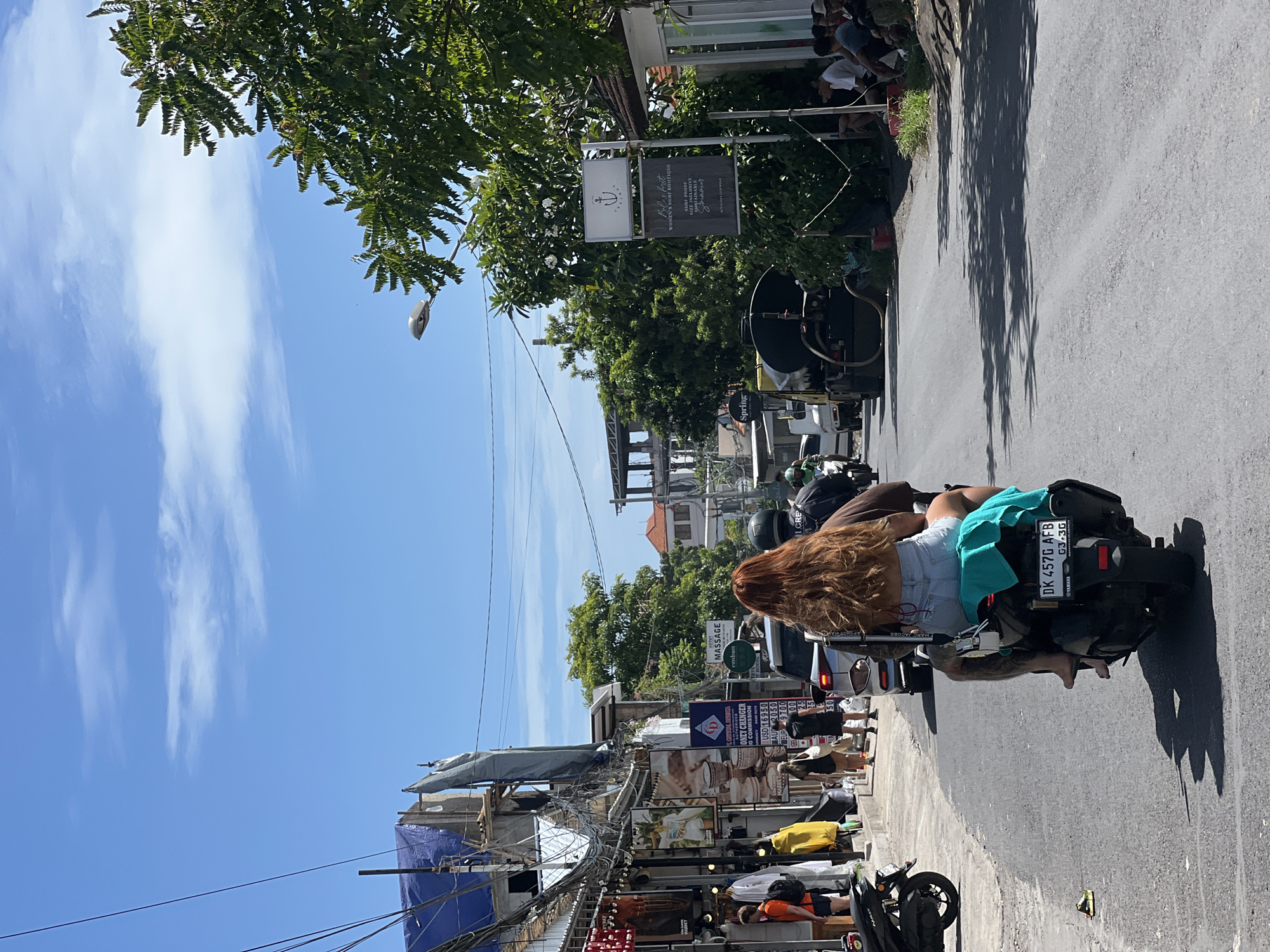 The image depicts a street scene in Kuta Selatan, Indonesia. A person with long, red hair is riding a black scooter, wearing shorts and a green top. The scooter's license plate reads "DK 457G AFB". The street is lined with shops and businesses, including a money changer and a women's surf boutique. There are several parked scooters and pedestrians on the sidewalks. The sky is blue with scattered clouds. Overhead power lines are visible.