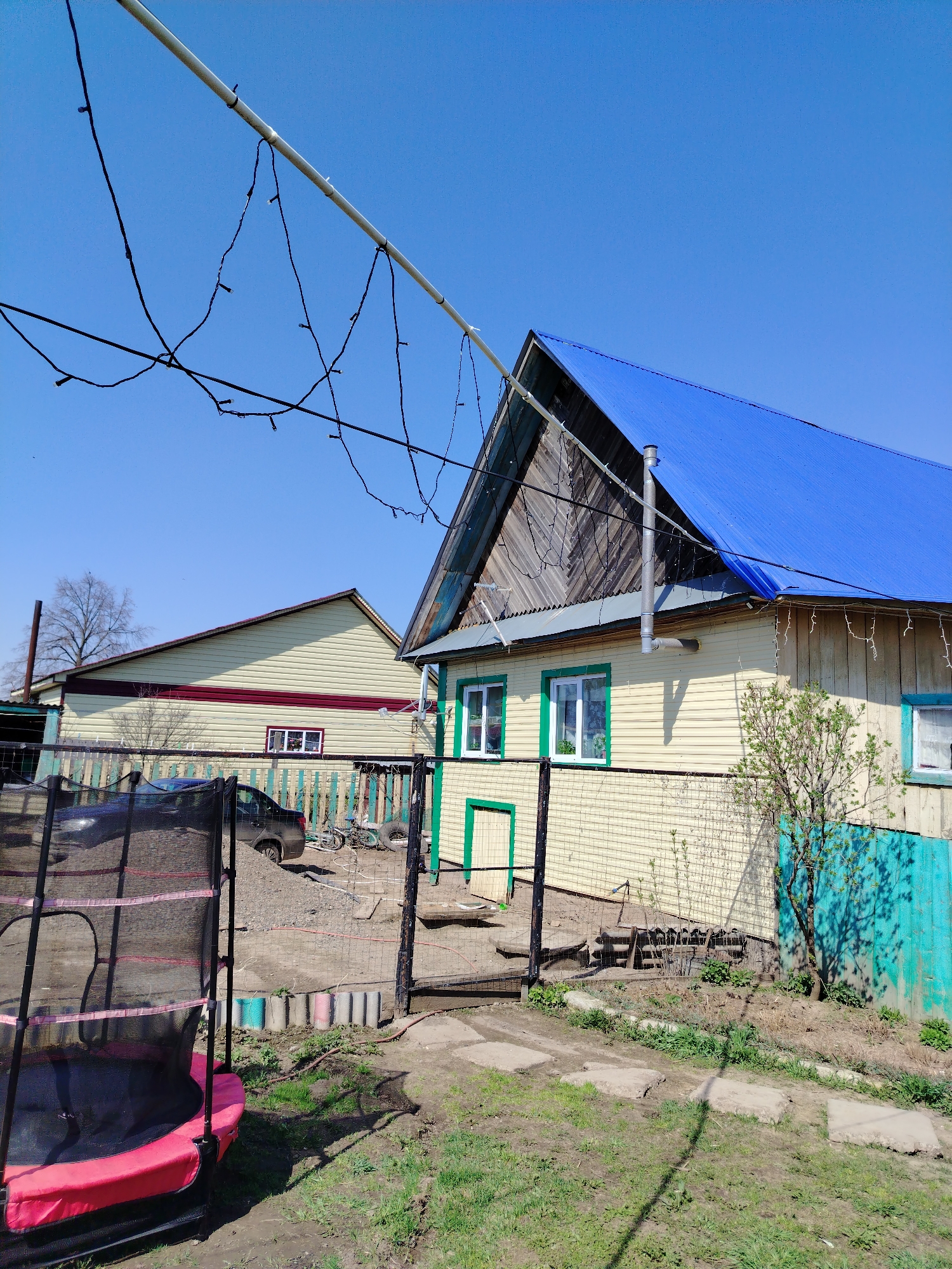 The image shows a residential scene in Гожан, Russia. A yellow, single-story house with a bright blue roof is in the foreground. The house has green trim around its windows and door. An external metal chimney is visible on the side of the house.
A black metal gate opens into a dirt yard with a few scattered concrete slabs leading to the house. To the left of the gate, a small trampoline with a red pad is partially visible. Beyond the gate and a wire fence, there is a pile of gravel, a car, and a bicycle. In the background, another yellow building with a red stripe is visible.
A white pole runs across the top of the frame, with black electrical wires and small lights strung along it. The sky is clear and blue. The overall impression is of a modest, rural or suburban dwelling in Гожан, Russia.