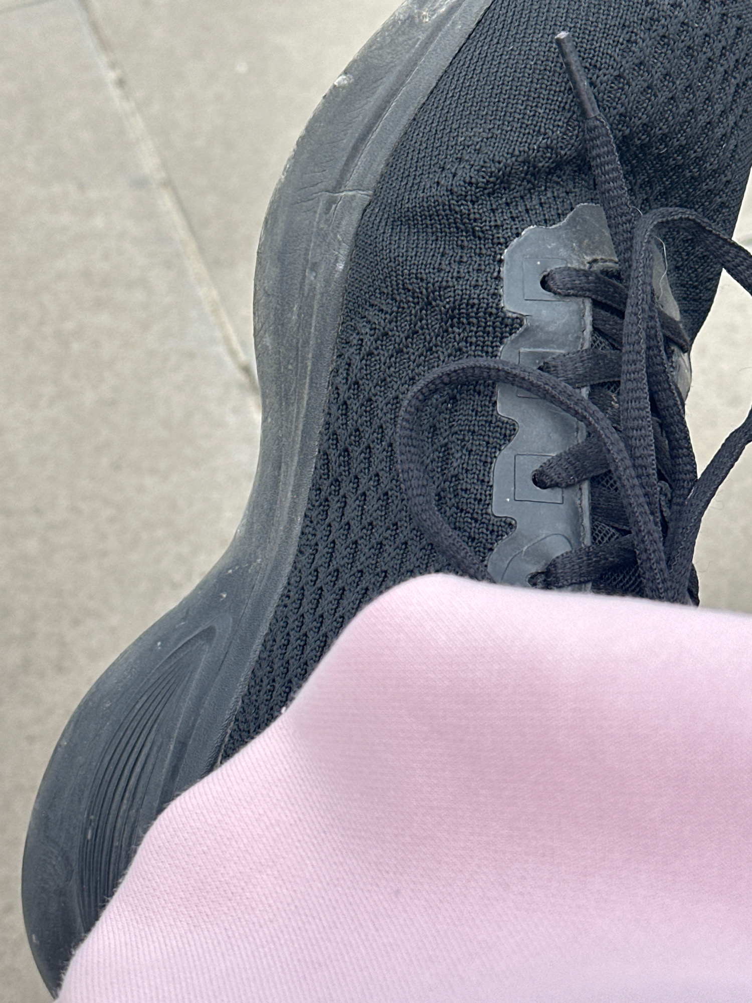 A close-up shot of a person wearing a black athletic shoe and light pink socks, standing on a sidewalk. The shoe is a dark color with black laces and a textured knit upper. A portion of the person's leg, covered by the pink socks, is visible in the lower right corner. The ground appears to be concrete paving stones. The scene depicts an ordinary urban setting, likely in Gyumri, Armenia, given the context provided.