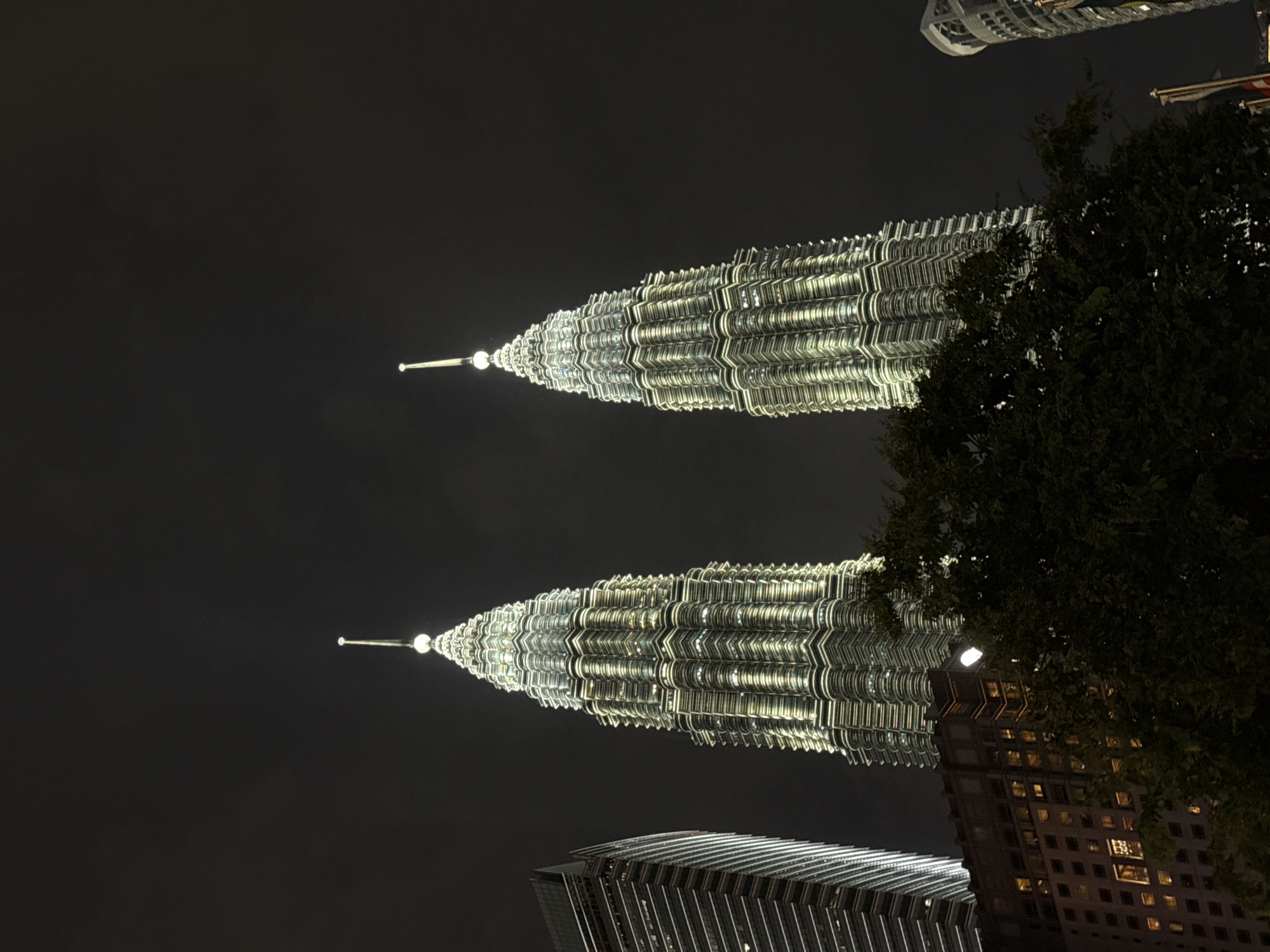 The image shows the Petronas Twin Towers in Kuala Lumpur, Malaysia, at night. The towers are brightly illuminated and dominate the skyline. A building with lit windows sits at the bottom of the frame, along with a tree with dark foliage partially obstructing the view of one of the towers.