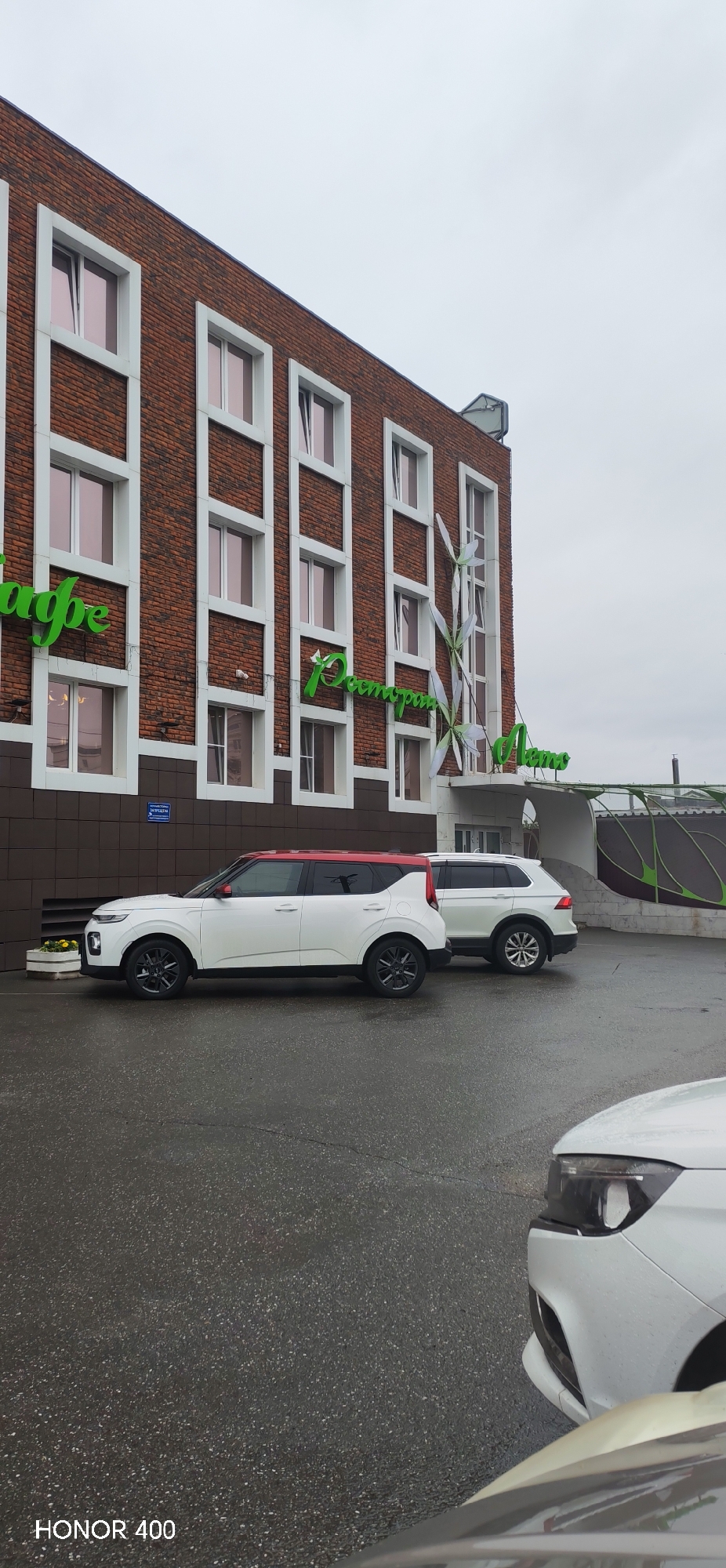The image depicts the exterior of a modern building, likely a cafe or restaurant, in Izhevsk, Russia. The building features a brick facade with white-trimmed windows arranged vertically in groups of three. Green neon signs, one reading "Кафе" (Cafe) and another "Ресторан" (Restaurant), are visible on the building's exterior. Adjacent to the building, a white SUV with a red roof and a white SUV are parked on a wet asphalt surface. In the foreground, the hood and headlights of a white car are visible, suggesting the photo was taken from within another vehicle or from a low vantage point. The sky is overcast, indicating a cloudy or possibly rainy day. The overall impression is of a commercial establishment on a typical day, with vehicles parked outside. The text "HONOR 400" is visible in the bottom left corner of the image.