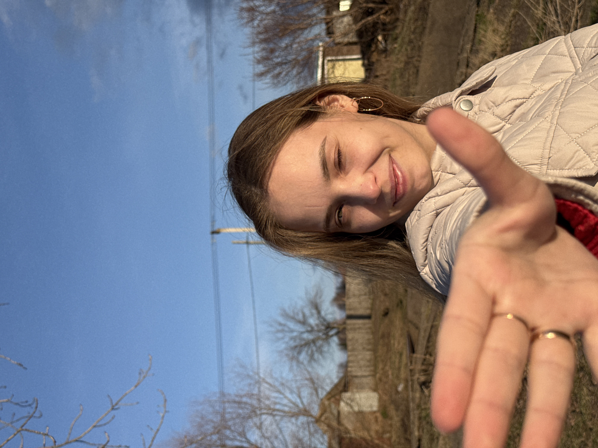 A brightly lit, tilted selfie-style portrait features a young person, likely female, smiling warmly with her eyes partially closed, possibly from direct sunlight. She has light brown hair and wears a light-colored, possibly beige or cream, quilted jacket over a red inner layer. Small gold hoop earrings are visible on her left ear. Her right hand is extended towards the camera in the foreground, slightly out of focus, showing two gold-toned rings on her ring and middle fingers.
The background consists predominantly of a clear, bright blue sky on the left side. The right and bottom portions reveal a dry, earthy landscape with bare deciduous trees, suggesting late autumn, winter, or early spring. Faint outlines of distant buildings or structures are visible, along with power lines running across the upper part of the frame. The strong sunlight indicates it is daytime, likely morning or afternoon, with good weather. The setting appears to be an outdoor, semi-rural or village area, consistent with a location like Chaikovsky, Russia. The person's expression conveys joy or playfulness. There is no visible text in the image.