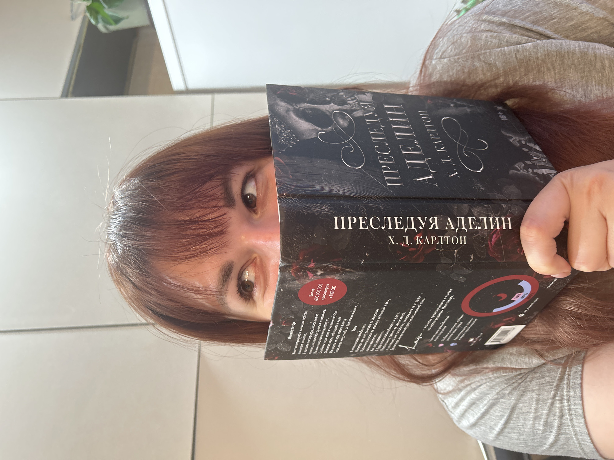 A person with auburn hair and bangs is holding the book "PRESLEDUYA ADELIN" (Pursuing Adeline) by H.D. Carlton in front of their face. The book is black with silver lettering. The person's eyes are visible above the book. They are wearing a gray shirt. The background includes a light-colored wall and a glimpse of a plant. The OCR text indicates the book is popular on TikTok and has received over 600,000,000 views. The location is not discernible as Фоки, Russia, but rather an indoor location.