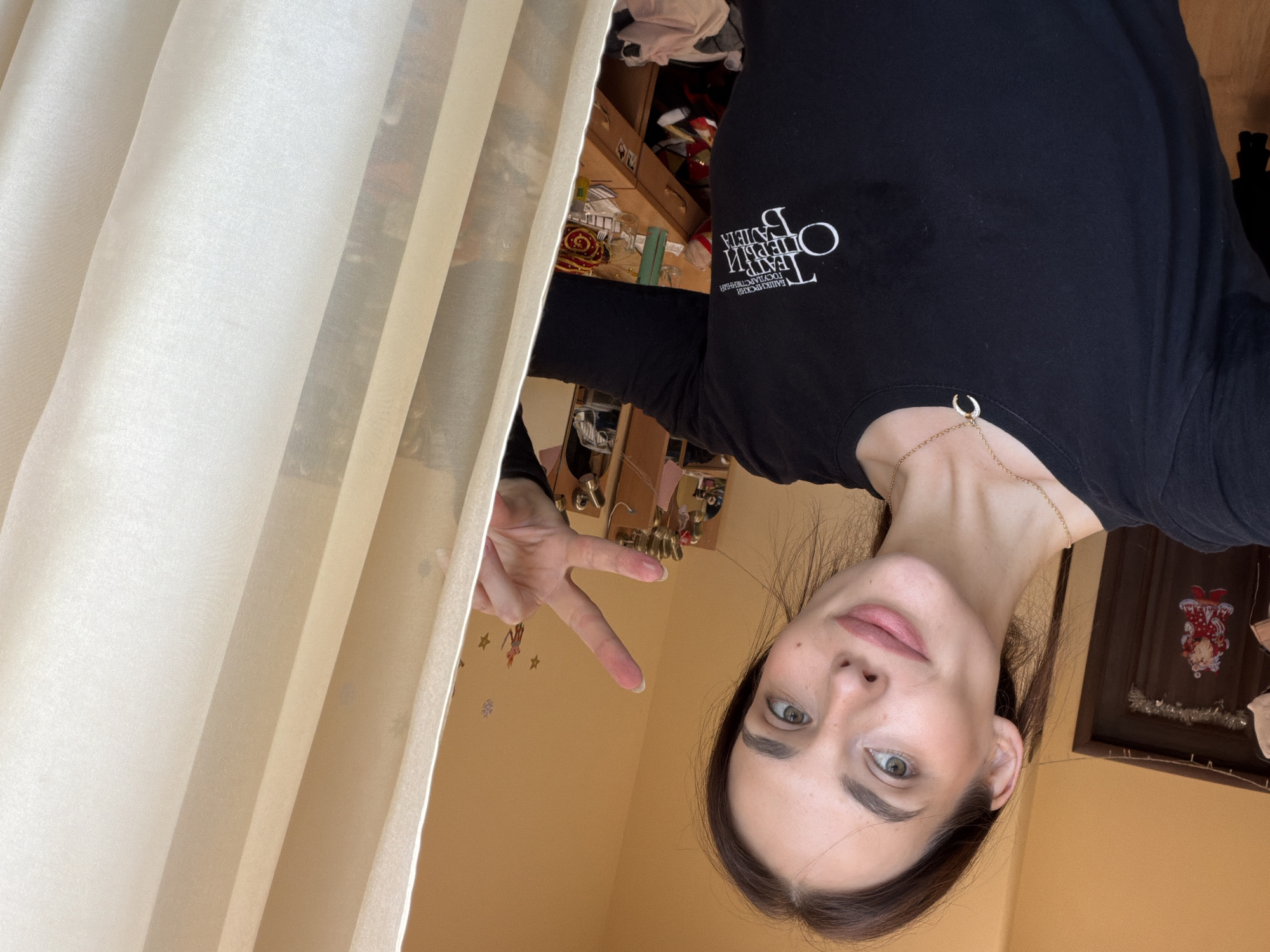 A young woman with dark hair, fair skin, and light green eyes takes a selfie in a room with yellow walls. She is wearing a black long-sleeved shirt with a white logo that reads "ТЕАТР ОПЕРЫ И БАЛЕТА" (Theater of Opera and Ballet) and a gold necklace with a crescent moon pendant. She is making a peace sign with her right hand, which has pink makeup on her fingers. To her left, a door is decorated with a picture of Santa Claus and tinsel. Behind her, a wooden desk is visible, cluttered with various items. A cream-colored curtain is to her right. The Bashkir State Opera and Ballet Theater is located in Ufa, Russia.