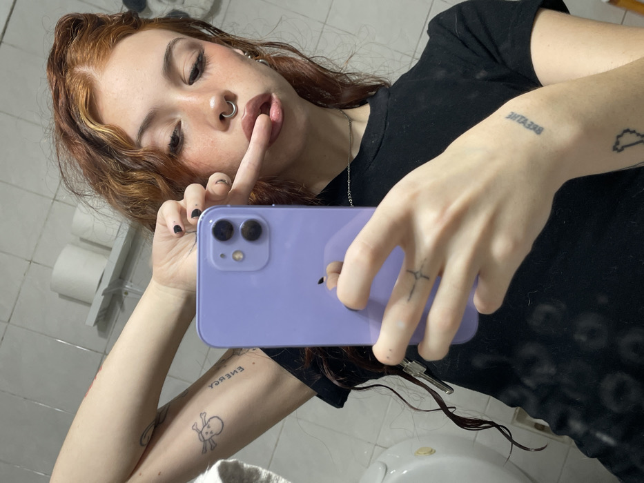 A red-haired woman holds a lavender smartphone while posing in a bathroom mirror. She has a nose ring and a finger to her lips in a shushing gesture. Black nail polish adorns her fingers. Tattoos on her arms read "ENERGY" and "BREATHE," with a skull and crossbones and a stylized state outline. The background reveals a toilet and toilet paper dispenser.