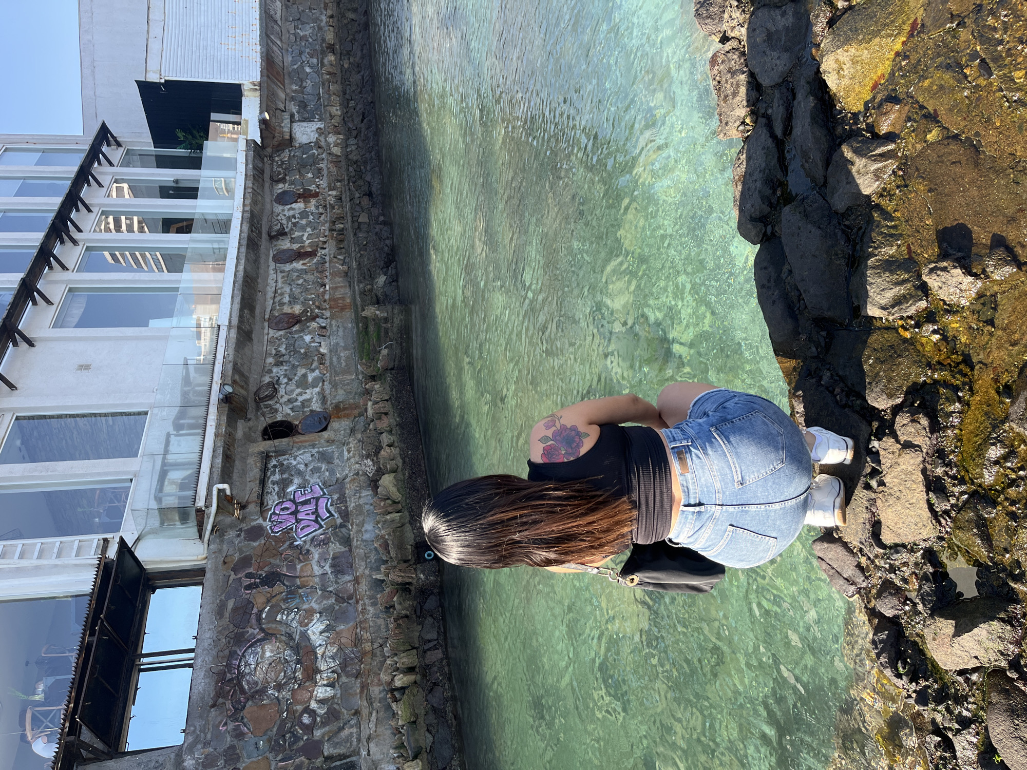 A person is kneeling on a rocky outcropping next to a body of water. The person has long dark hair, is wearing a black tank top and blue denim shorts, and has a tattoo on their upper back. They are positioned such that their back is facing the camera. The water is clear and appears shallow, with visible ripples and variations in color from light green to darker shades. The rocky outcropping is composed of dark-colored stones. To the left of the water is a building with a stone wall and a modern, multi-story section with large windows. There is graffiti on the stone wall. The sky is a clear, light blue. The location is Unknown, Unknown.