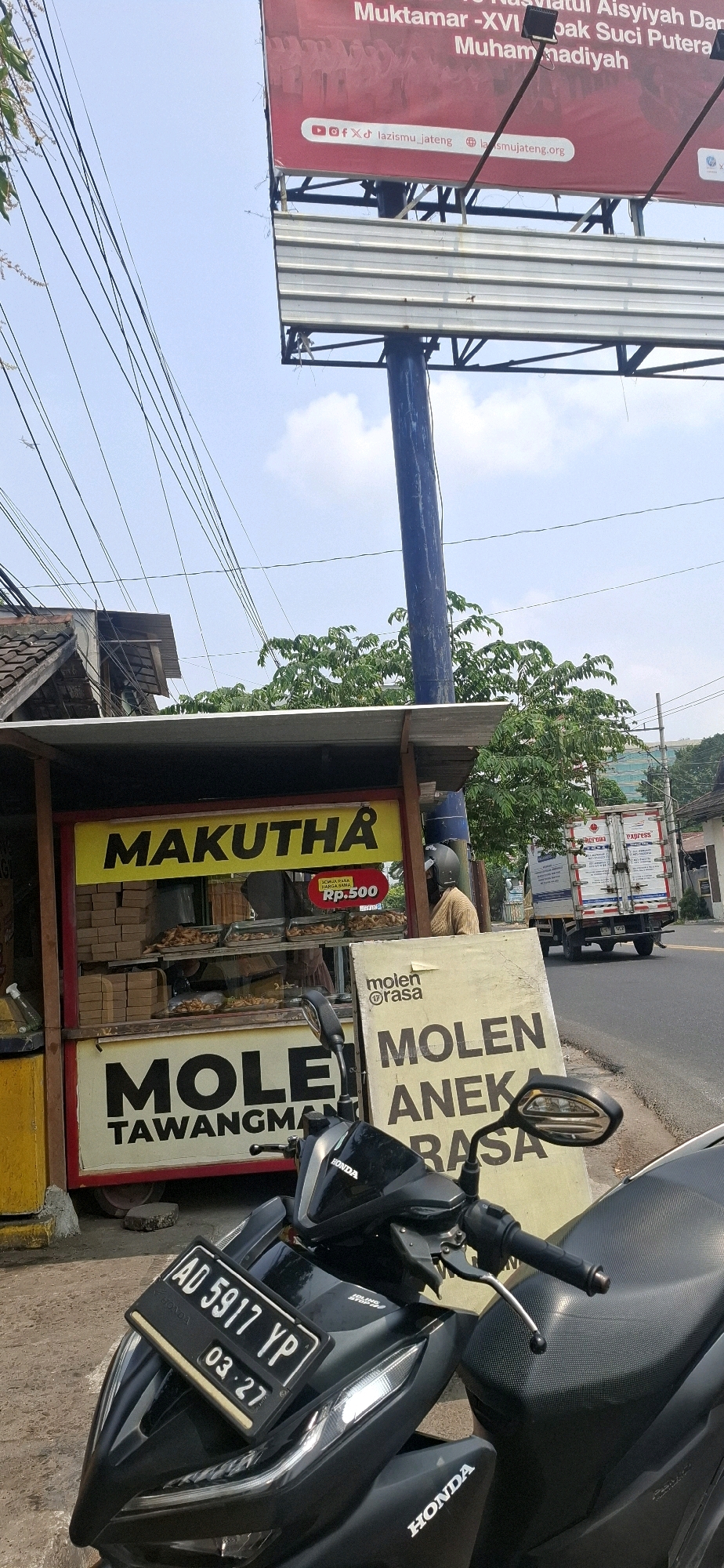 In Blulukan, Indonesia, a street scene unfolds under a bright sky with scattered clouds. The foreground is dominated by a black motorcycle, its license plate reading "AD 5917 YP" with an expiration date of "03.27". To the left of the motorcycle is a food stall with a yellow awning and a sign that reads "MAKUTHA" in large yellow letters on a red background. Below this, another sign proclaims "MOLEN TAWANGMAN" in large black letters. Inside the stall, trays of fried snacks are displayed. Adjacent to the food stall is a light yellow sign with the words "molen rasa" and "MOLEN ANEKA RASA" in large black letters. In the background, a white truck with "EXPRESS" written on its side drives down a paved road. Overhead, numerous electrical wires crisscross the sky, and beyond the road, trees and buildings are visible, including a multi-story building in the distance. A tall blue pole supports a large billboard displaying red text and social media icons, including "lazismu_jateng" and "lazmujateng.org". The overall atmosphere suggests a typical day in a bustling Indonesian town.