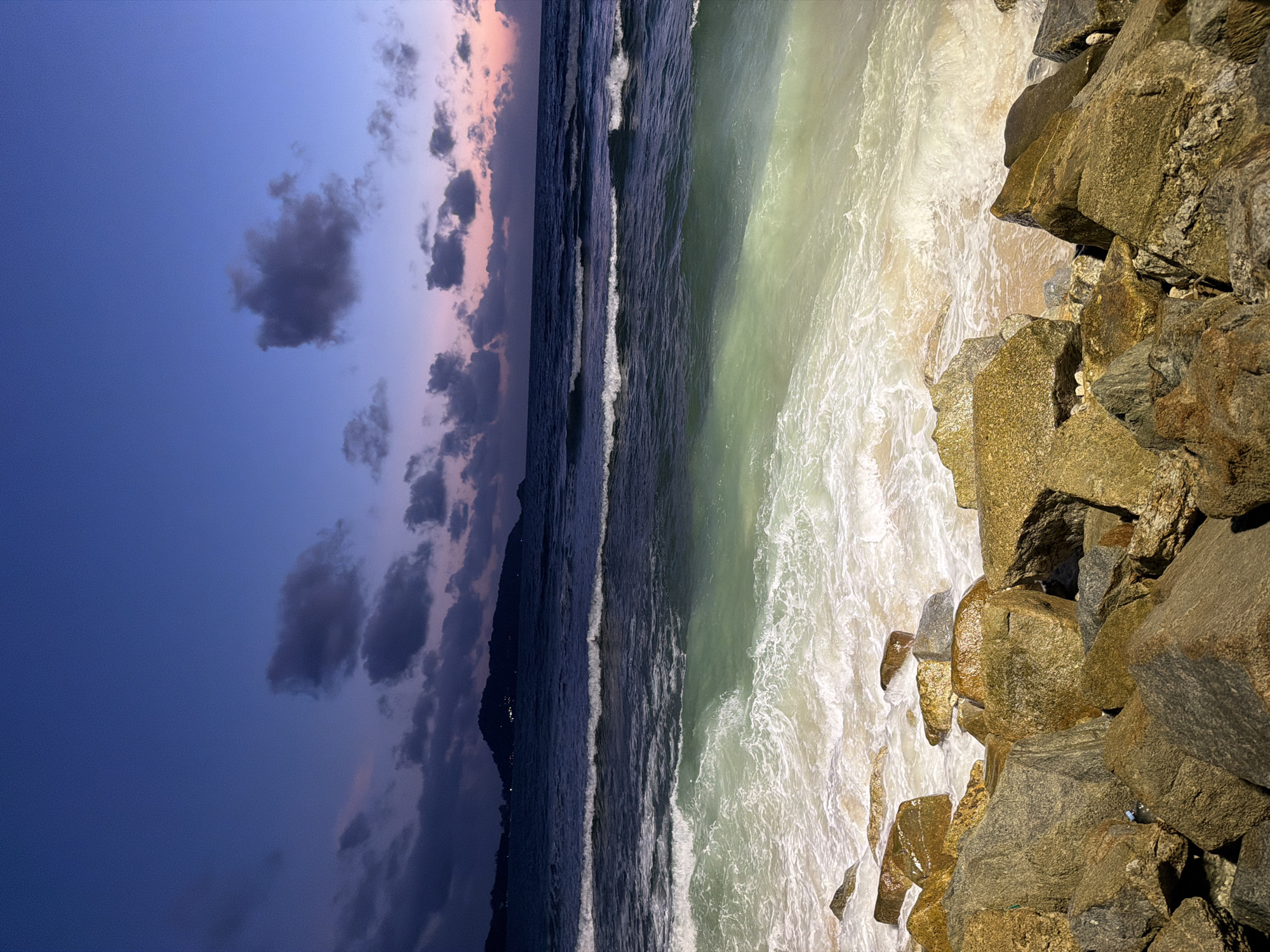This is a nighttime photograph of a rocky shoreline in Patong, Thailand. The sky is a deep blue with streaks of purple and pink from the setting sun. Clouds are scattered across the sky, some dark and heavy, others lighter and wispy. The ocean is dark and choppy, with waves breaking on the shore. In the foreground, large rocks are piled up along the beach. A distant hill with lights is visible on the horizon.