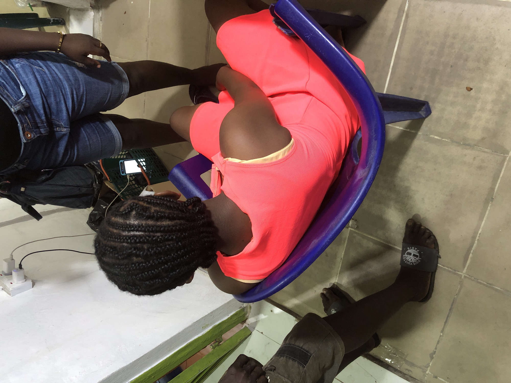 A dorsal view shows a Black person sitting in a blue plastic chair, facing away from the camera. The person has braided hair and is wearing a bright pink sleeveless dress with a yellow lining visible at the neckline. Their legs are visible, and they are wearing dark-colored flip-flops. In the foreground to the left, a person's legs and feet are visible, wearing grey shorts and black flip-flops. The environment appears to be indoors, with tiled flooring and a white wall. A power outlet with two plugs is visible on the wall. A green-painted door frame is partially visible on the left. In the background, a green plastic basket containing a mobile phone is visible, along with a black duffel bag and other dark objects. The scene is located in Ado, Nigeria.