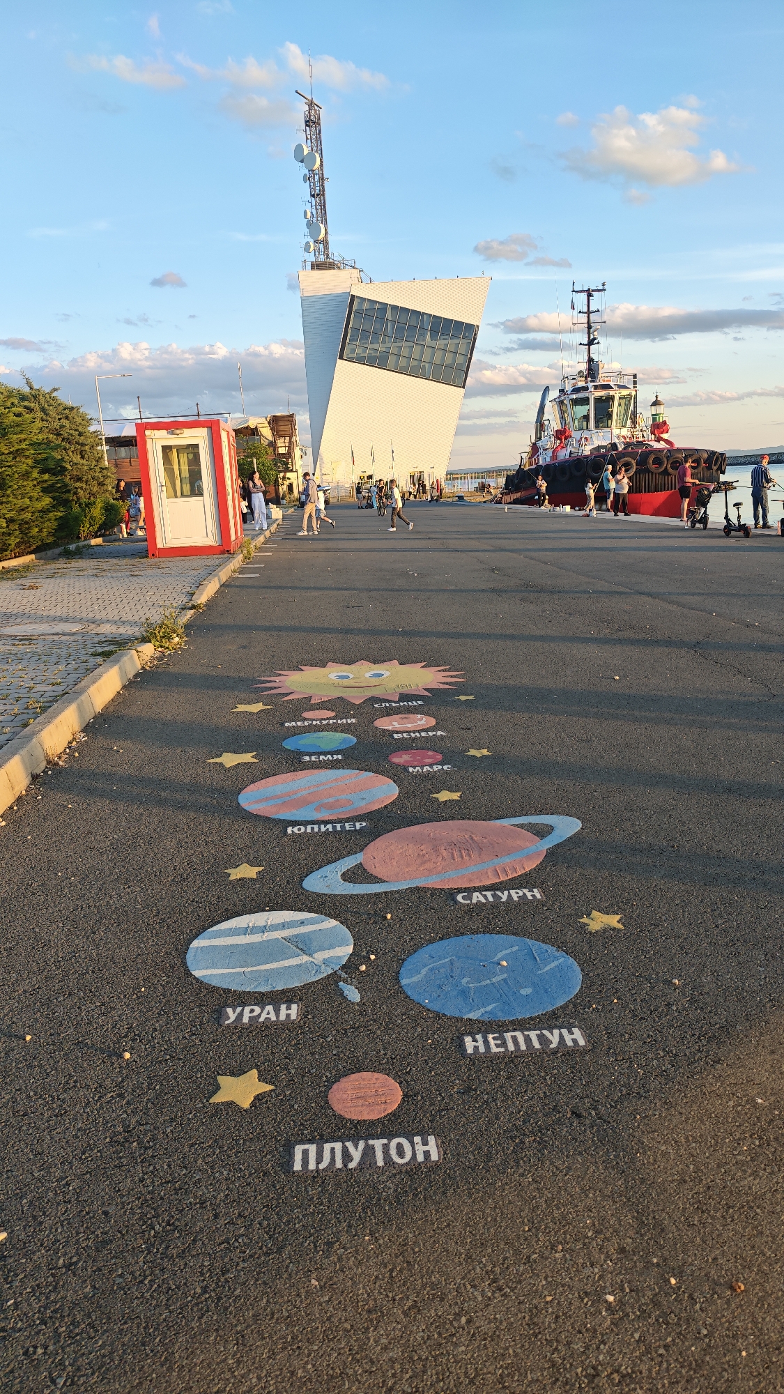 The image shows a street painted with a depiction of the solar system, located in Burgas, Bulgaria. From the foreground to the background, the solar system is illustrated, with the "Sun" (Слънце) closest to the viewer, followed by "Mercury" (Меркурий), "Venus" (Венера), "Earth" (Земя), "Mars" (Марс), "Jupiter" (Юпитер), "Saturn" (Сатурн), "Uranus" (Уран), "Neptune" (Нептун), and "Pluto" (Плутон). Yellow stars are interspersed between the painted planets.
The street appears to be a public area near the port. In the background, there is a modern white building with a tall communication tower, and a red and white tugboat is docked nearby. People are seen walking and gathering around the port area.
Further to the left, a small red and white booth or shed can be observed. The sky is partly cloudy.