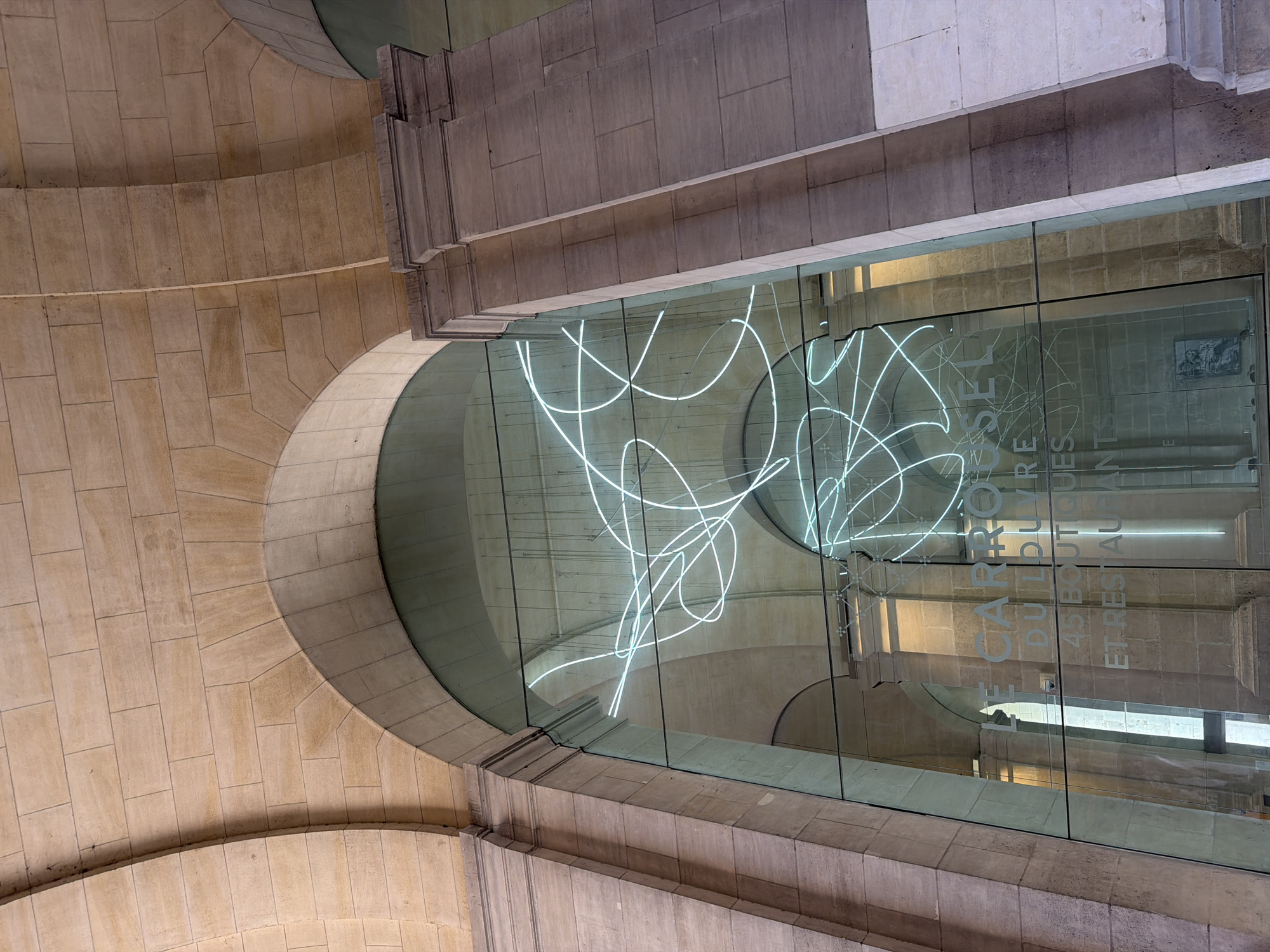 The image depicts a view looking up at a glass-walled structure in Paris, France. The structure is "LE CARROUSEL DU LOUVRE 45 BOUTIQUES ET RESTAURANTS" as shown on the glass walls. The glass wall reflects the architectural details of the building on the outside, including arches and stonework. There is a white abstract light sculpture inside the glass-walled structure. The scene appears to be taken from a slightly angled perspective, looking up towards the structure, showcasing the intersection of the modern glass architecture with the classic stone building elements.