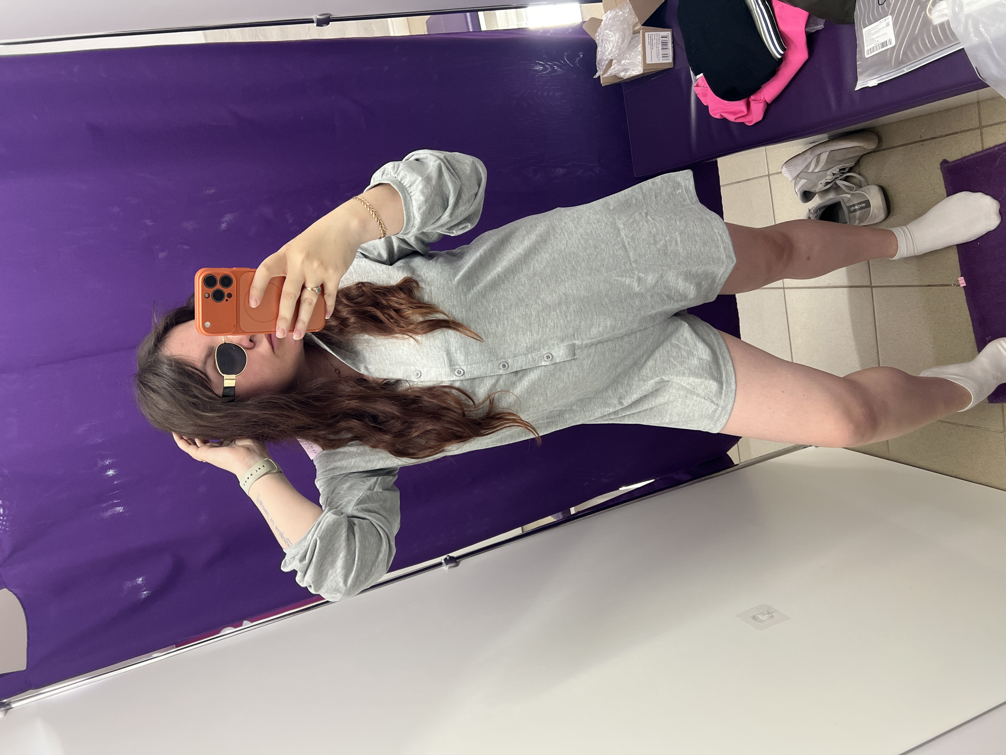 A woman stands in a fitting room, taking a selfie in a mirror. She is wearing a light gray, long-sleeved romper with a collared neckline and button detailing down the front. She has dark, wavy hair and is wearing gold-rimmed sunglasses. Her right hand is raised, touching her hair, while her left hand holds an orange smartphone, which is capturing the image. She is also wearing a green smartwatch on her left wrist and a gold bracelet on her right wrist, along with a ring on her right ring finger. On her feet, she has white socks and gray sneakers. The fitting room has a large purple curtain in the background and white walls. To the right, there is a purple bench with some clothing items and a cardboard box. The floor is tiled. The location is not specified to be in Noviy, Russia.