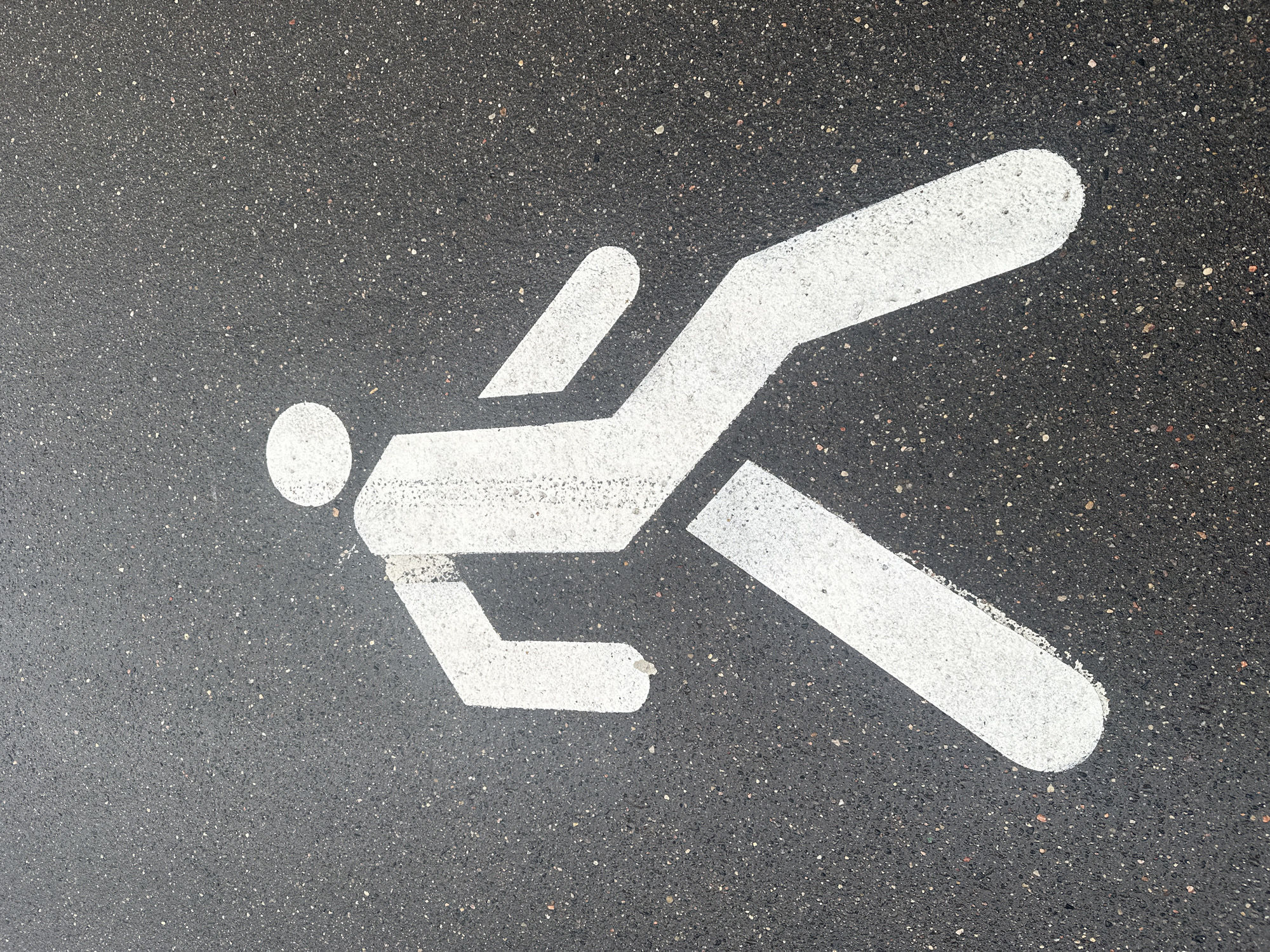 A white pedestrian crossing symbol is painted on a dark asphalt surface. The symbol depicts a stylized human figure walking, with a circular head, rectangular torso, and arms and legs extended as if in mid-stride. The asphalt surface is coarse-grained and appears slightly damp, with small light-colored aggregate particles visible throughout. The lighting suggests an overcast day. This image was taken on May 13, 2023, in Minsk, Belarus.