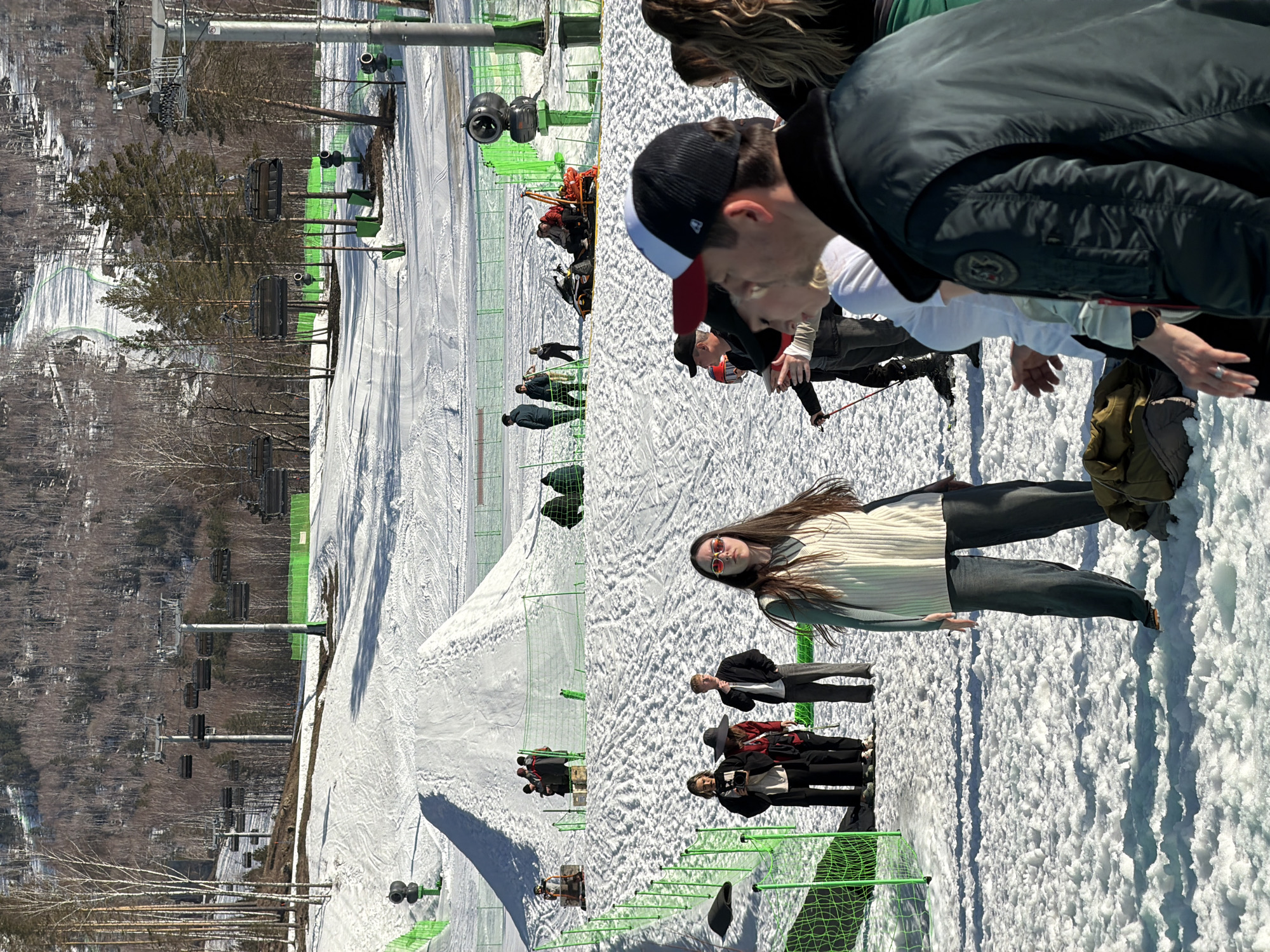 The image shows a snowy ski resort in Озёрное, Russia. Several people stand on the snow. Some are near a green fence, others near a ski lift. One person with long brown hair wears sunglasses and stands prominently in the center. Some people are skiing or snowboarding. The background includes a hillside covered with trees. The sky is bright.