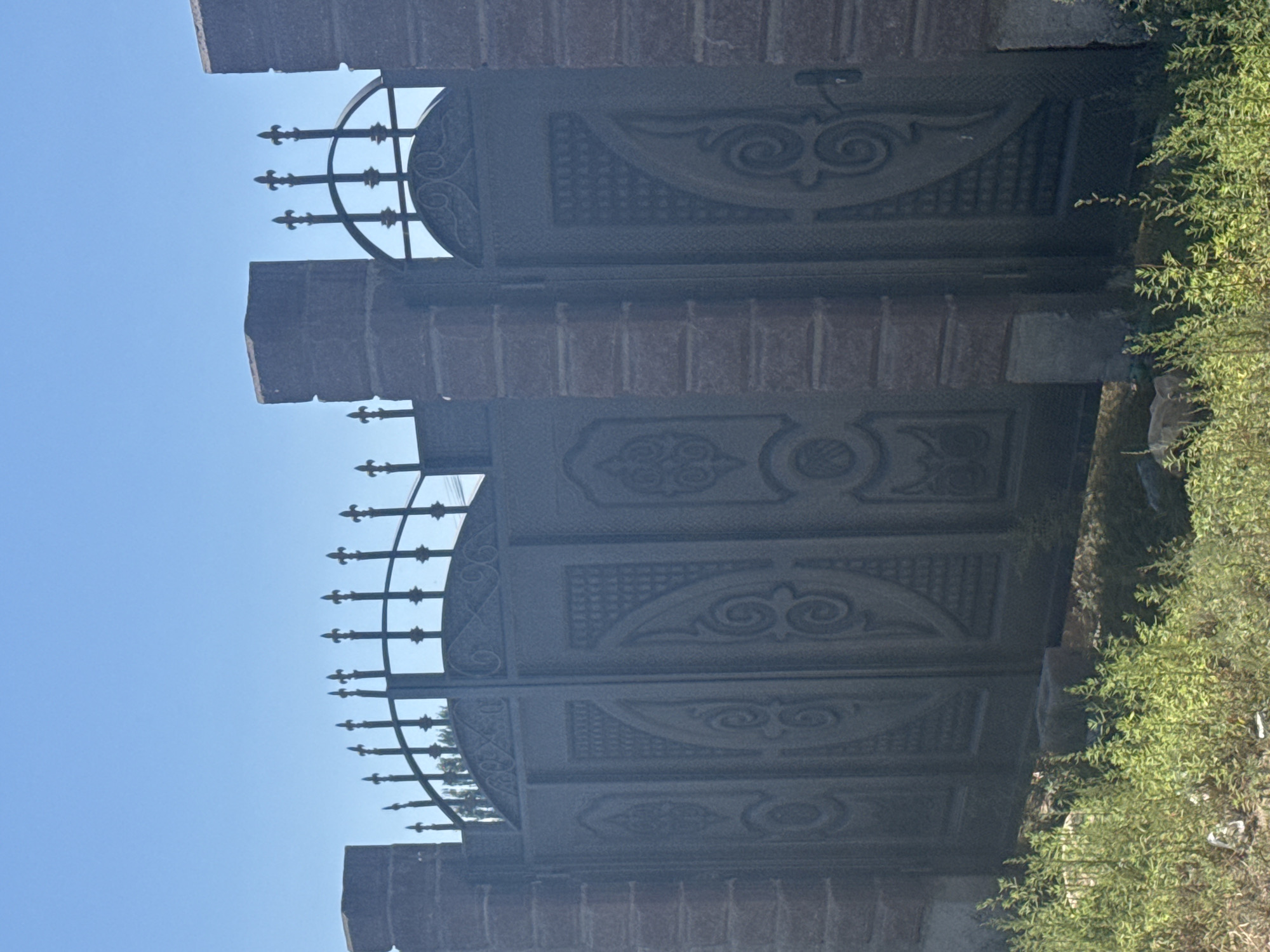 The image shows a large, ornate, dark metal double gate set between two brick pillars. The gate is decorated with intricate patterns, including swirling designs and what appear to be decorative reliefs. Above the gate, a series of pointed finials are visible, also made of metal. The sky is clear and blue, suggesting a sunny day. In the foreground, there is overgrown green vegetation. The scene appears to be outdoors, possibly in a residential or private property area. The location is identified as Sary-Oy, Kyrgyzstan.