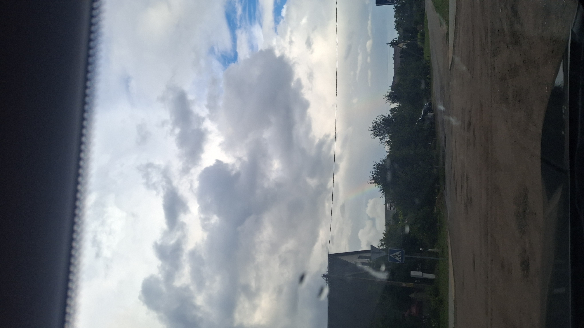 The image depicts a cloudy sky with a faint rainbow visible in the distance. In the foreground, a dirt road with numerous potholes leads towards a cluster of trees and houses. A pedestrian crossing sign is visible on the left side of the road, near a dark-colored house. The scene appears to be a rural or suburban area in Stowbtsy, Belarus. The viewpoint is from inside a vehicle, as evidenced by the reflection on the windshield.
