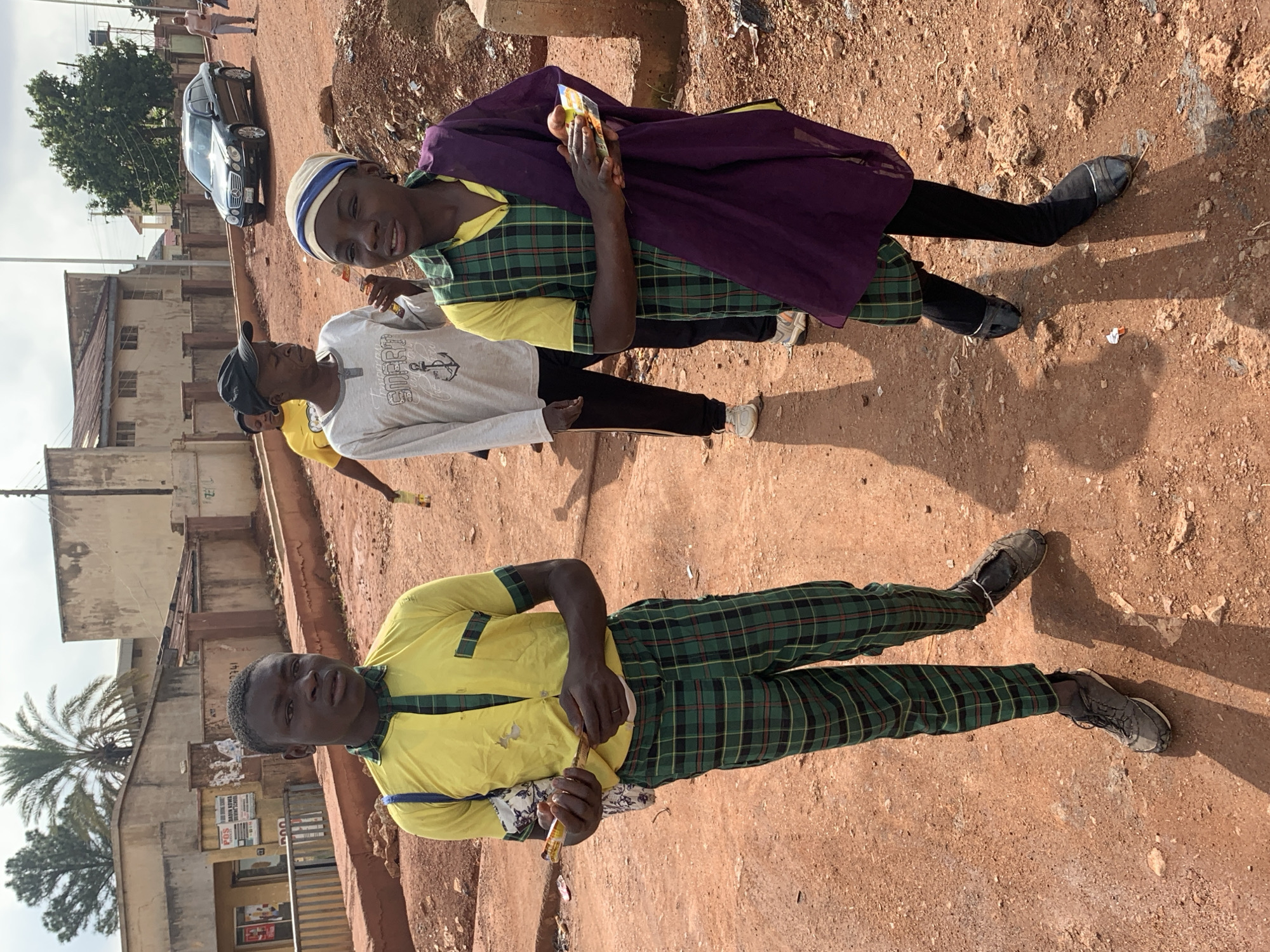 The image captures a group of young people in Gura Top, Nigeria, seemingly engaged in a casual outdoor gathering. Two young men are prominently featured, both wearing plaid school uniforms. One, in the foreground, sports a yellow short-sleeved shirt with green plaid accents and green plaid trousers. He is holding a small object, possibly a snack or a toy. The other young man, standing slightly behind and to the left, wears a light-colored long-sleeved shirt with a grey plaid pattern and dark trousers. He has a blue and white headwrap and is looking towards the left of the frame. A third individual, partially visible behind them, appears to be wearing a purple garment over a green plaid shirt.
The setting is an unpaved, reddish-brown ground, typical of many outdoor areas in Nigeria. In the background, a black car is parked, and to the left, there are buildings with a slightly weathered appearance. A palm tree is visible in the distance, suggesting a tropical or subtropical climate. The lighting indicates it is daytime, likely late afternoon given the long shadows cast on the ground. The overall mood seems relaxed and informal, with the subjects appearing comfortable and at ease. There is no overt text visible in the image that provides additional context about the specific activity or event.