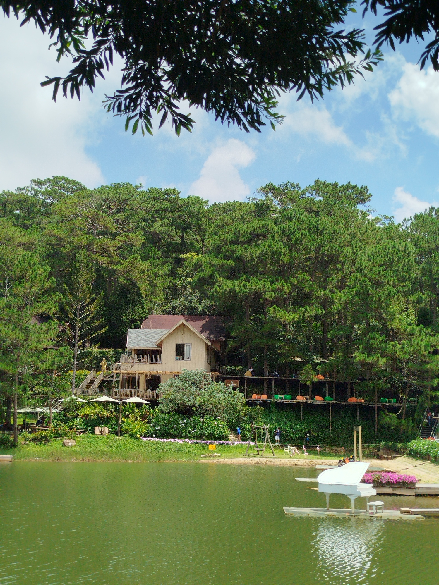 The image depicts a serene lakeside scene, likely located in Cam Ly, Da Lat, Vietnam. A tranquil green lake dominates the foreground, reflecting the lush, pine-covered hillsides that rise behind it. Nestled amongst the trees is a multi-level wooden house with a light-colored facade and a reddish-brown roof. Several balconies and outdoor seating areas are visible on the house. A set of wooden stairs leads up to the house from the lakeshore.
Along the water's edge, there is a grassy area with some seating and umbrellas, suggesting a recreational or dining space. Further to the right, a unique feature is present: a white grand piano is set up on a floating platform in the lake, accompanied by a small stool. The piano appears to be an artistic installation or an attraction. The background is filled with dense evergreen trees. The sky is partly cloudy, with patches of blue visible. Several people are scattered throughout the scene, engaged in various activities like walking and relaxing. Some are on the wooden platforms near the house, while others are near the lake's edge. The overall atmosphere is peaceful and picturesque.